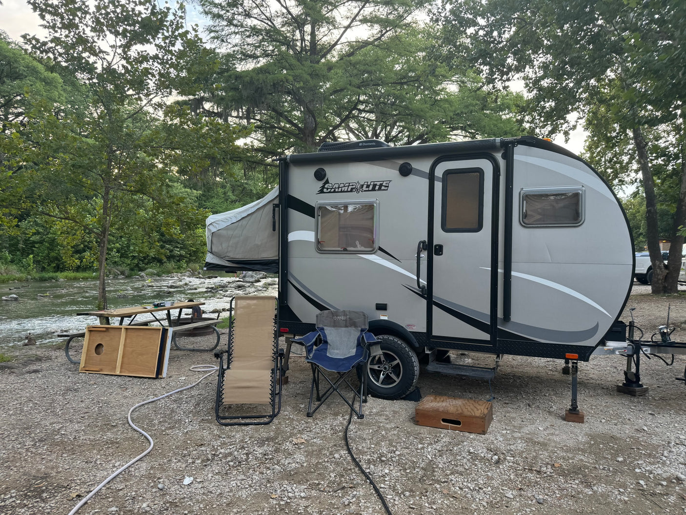 RV rental campsite by a river: a grey camper with a pop-out bed, picnic table, and chairs on gravel.