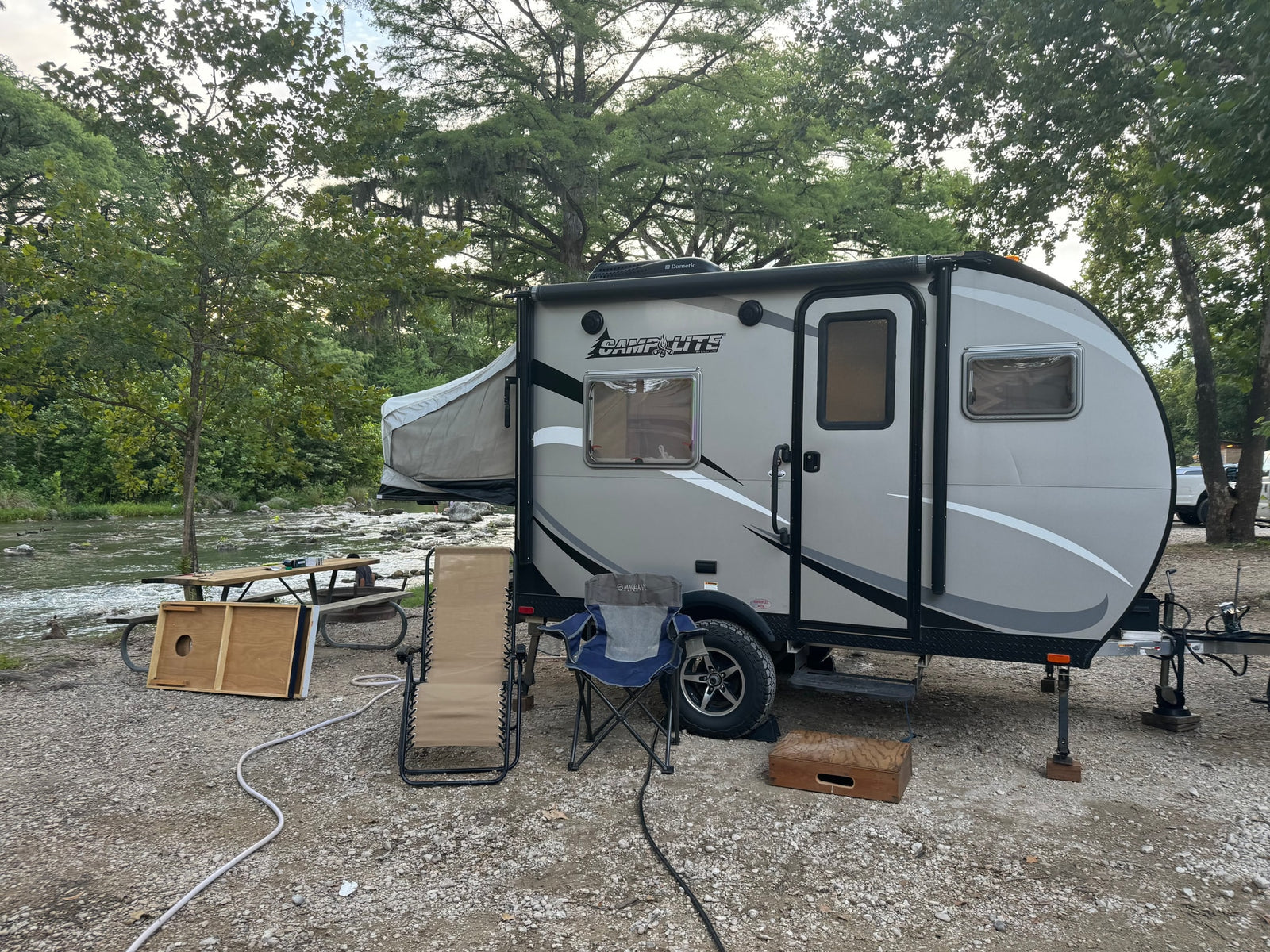 RV rental campsite by a river: a grey camper with a pop-out bed, picnic table, and chairs on gravel.
