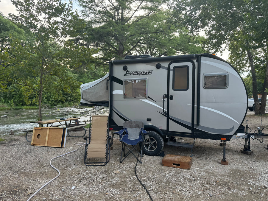 RV rental campsite by a river: a grey camper with a pop-out bed, picnic table, and chairs on gravel.