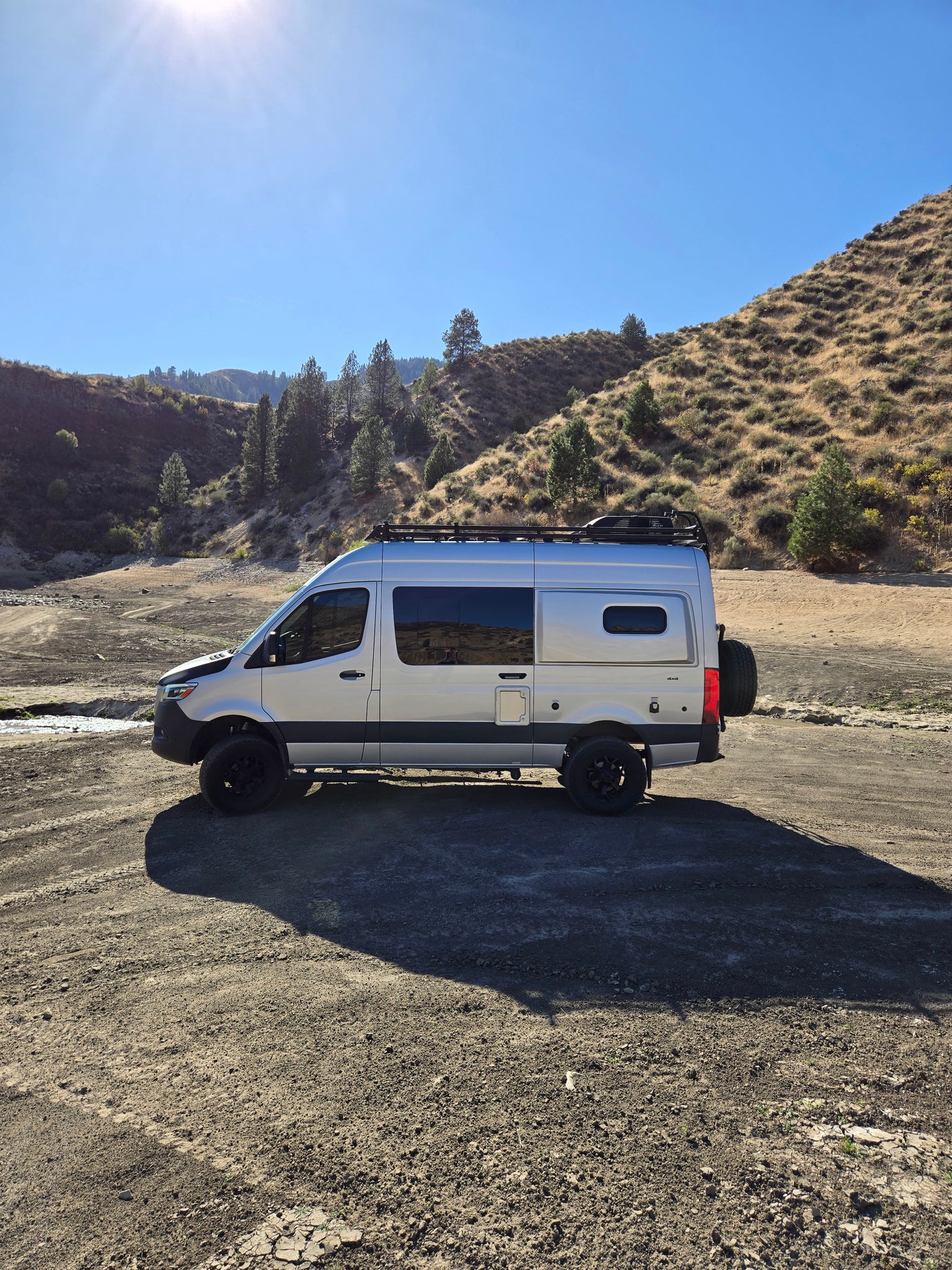 Explore nature in this adventure-ready RV rental! Silver camper van on a sunny dirt road by dry hillsides & a stream.