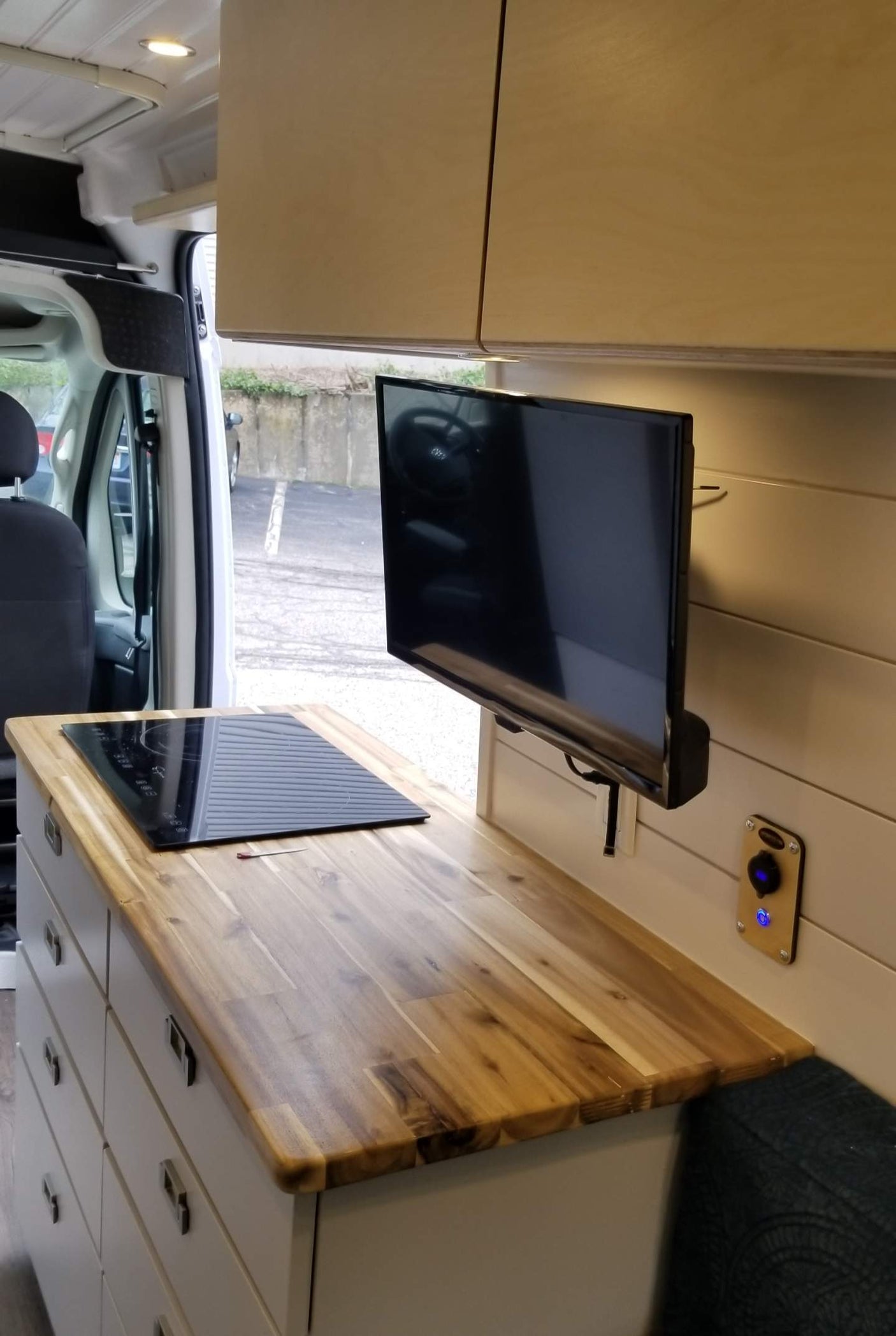 Inside a sleek RV rental van: a kitchen with wood counter, induction cooktop, drawers, and mounted TV. Compact living ready.