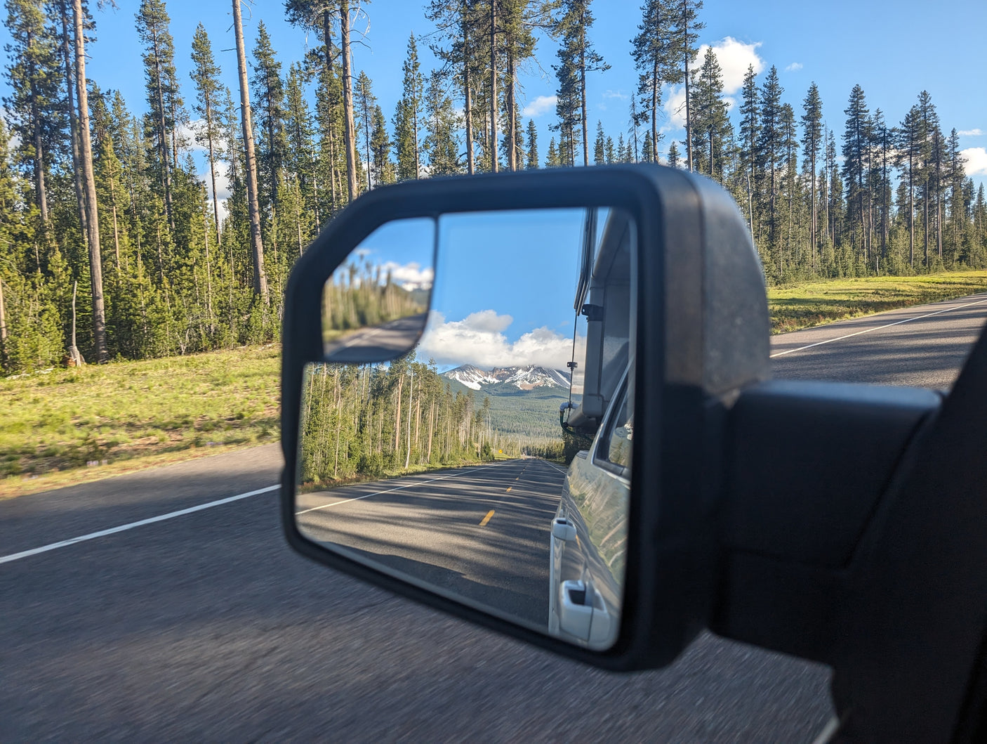 Scenic drive from an RV rental, showing road, pine forest, snowy mountains, and a side mirror reflection.