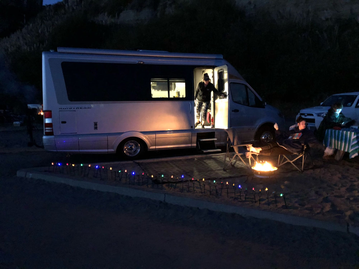 Enjoying an RV rental adventure: An Airstream campervan at night with people by a cozy campfire, illuminated for outdoor relaxation.