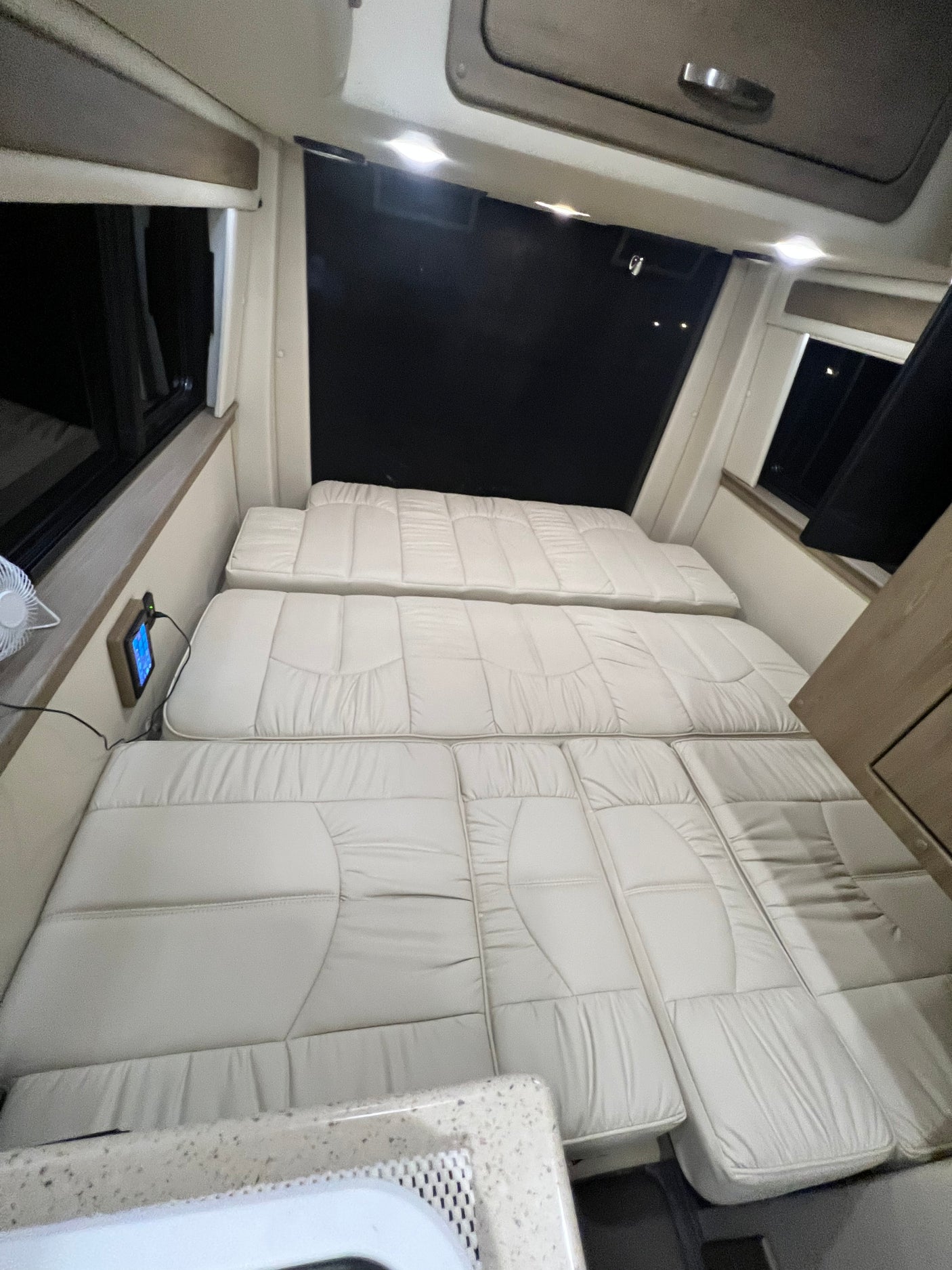 Spacious RV rental interior featuring light-colored cushions arranged as a bed, with windows & control panel access.