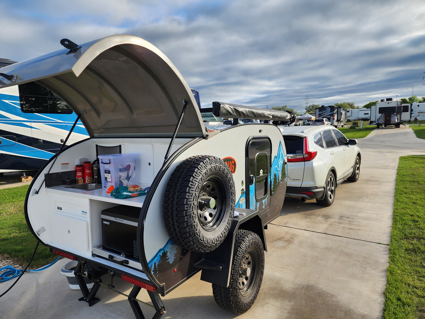 Compact rv rental teardrop camper with outdoor kitchen set up, hitched to SUV at an RV park.