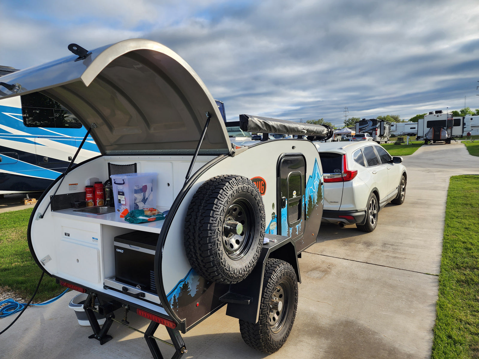 Compact rv rental teardrop camper with outdoor kitchen set up, hitched to SUV at an RV park.