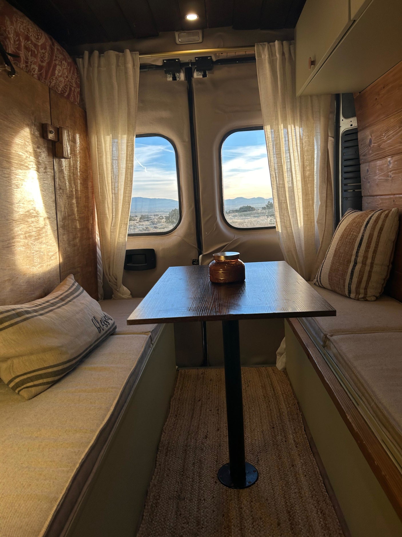 Cozy RV rental interior features a dining area with scenic mountain views, wood paneling, soft curtains, and cushions.
