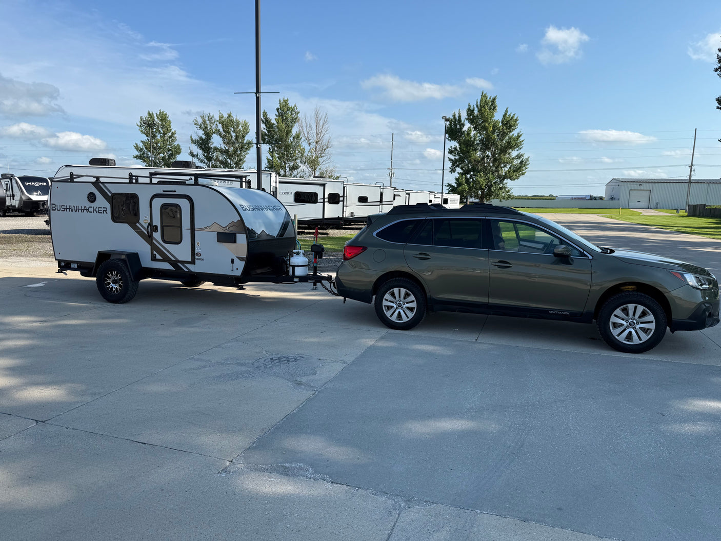 A dark green Subaru Outback tows a compact Bushwhacker rv rental camping trailer in a sunny outdoor lot with other RVs.