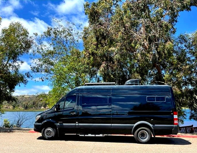This black RV rental van, parked by a calm lake with trees and a blue sky, offers outdoor adventure.