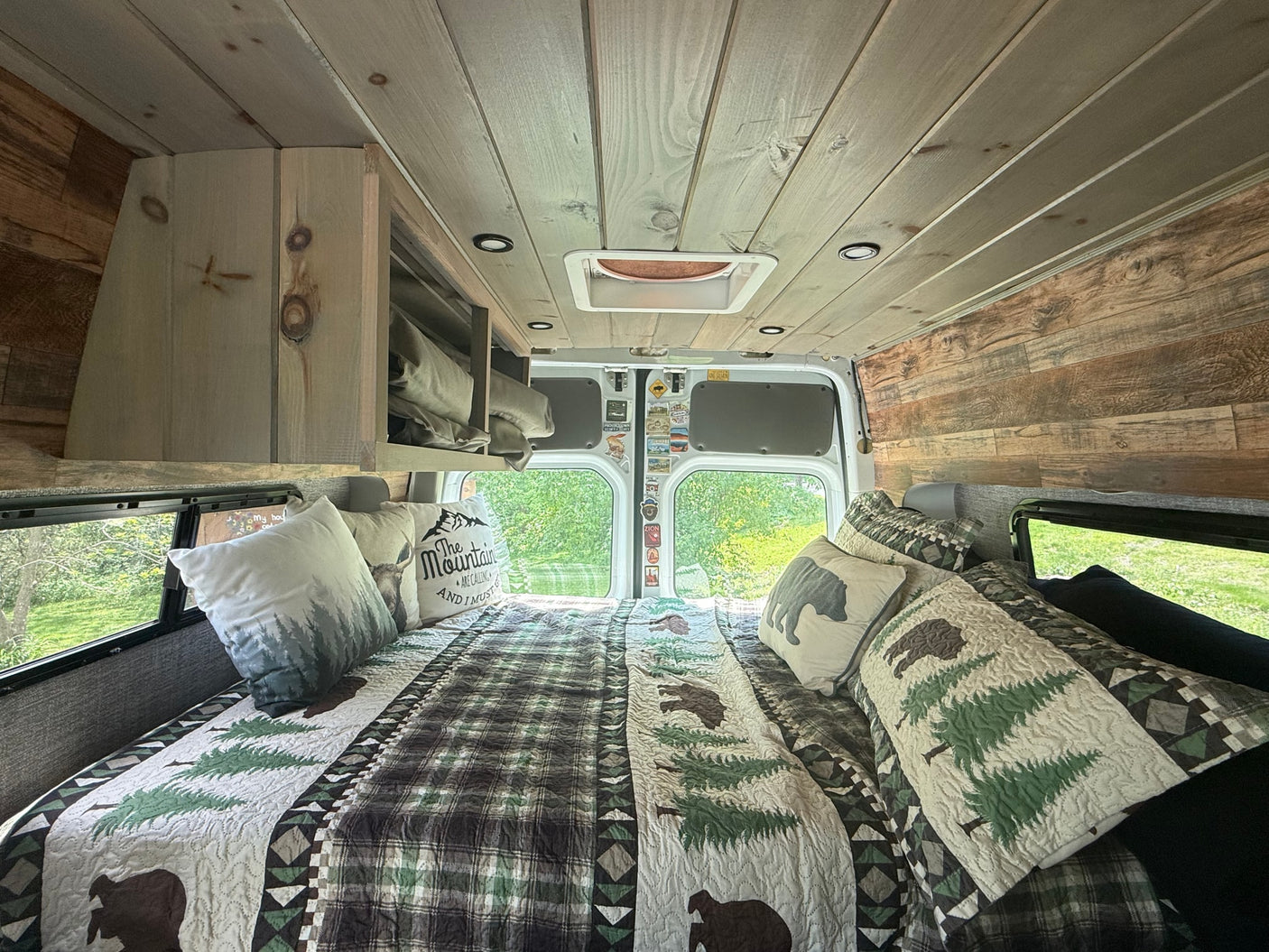 Cozy RV rental camper van bedroom with rustic wood paneling, woodland-themed bedding, and scenic windows.