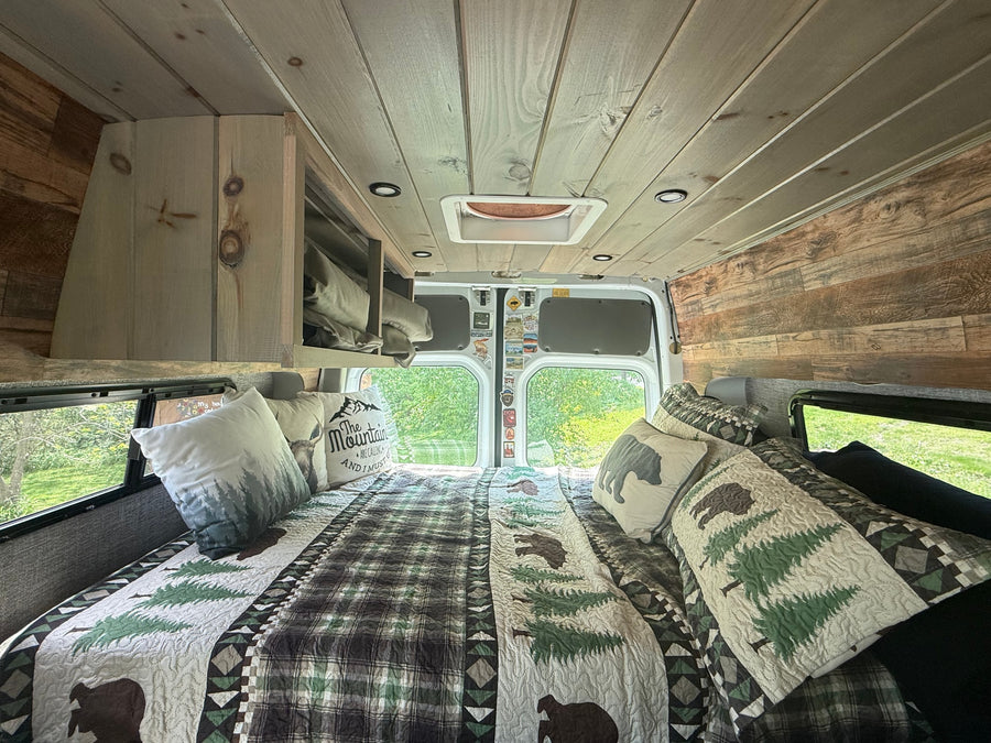 Cozy RV rental camper van bedroom with rustic wood paneling, woodland-themed bedding, and scenic windows.