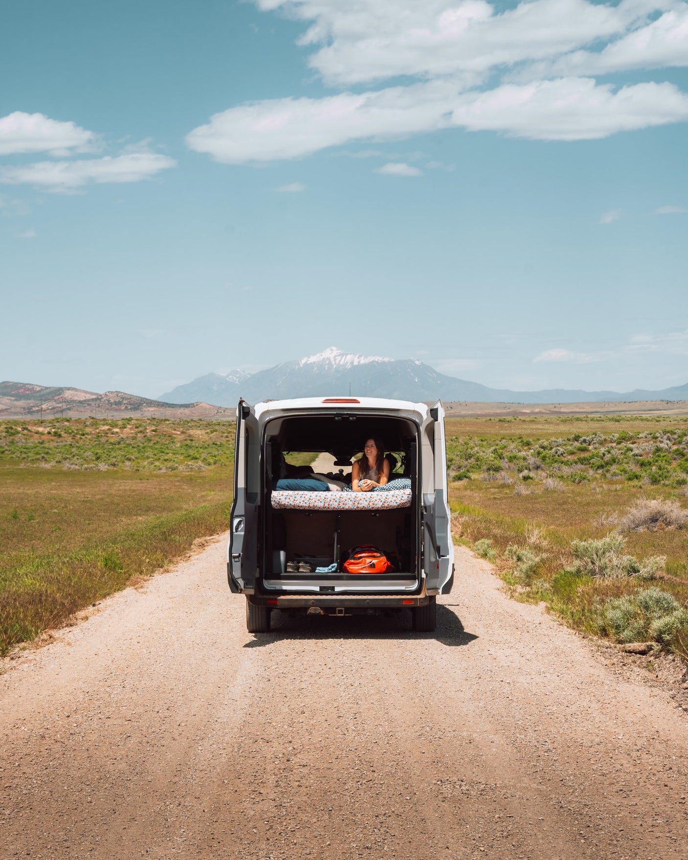 A woman relaxes in an RV rental van's open back, enjoying mountain views from her bed on a remote dirt road.