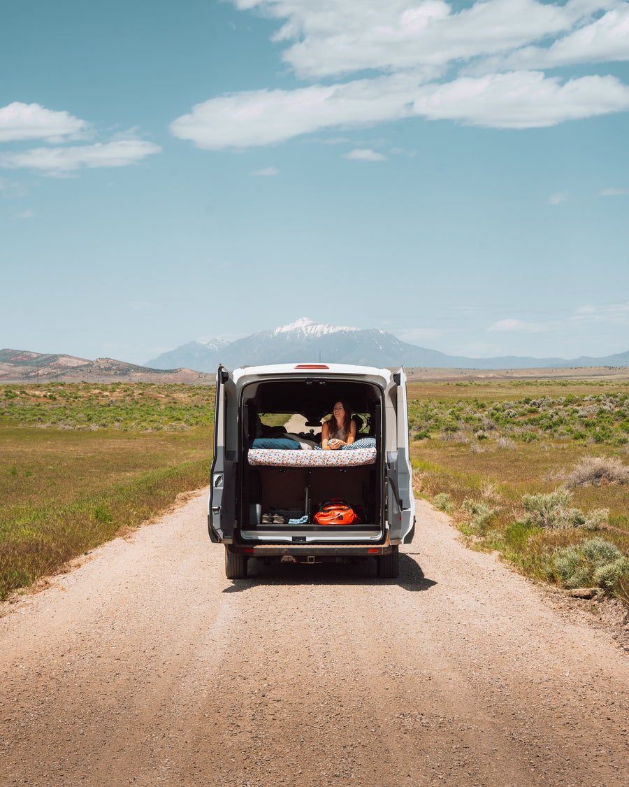 A woman relaxes in an RV rental van's open back, enjoying mountain views from her bed on a remote dirt road.
