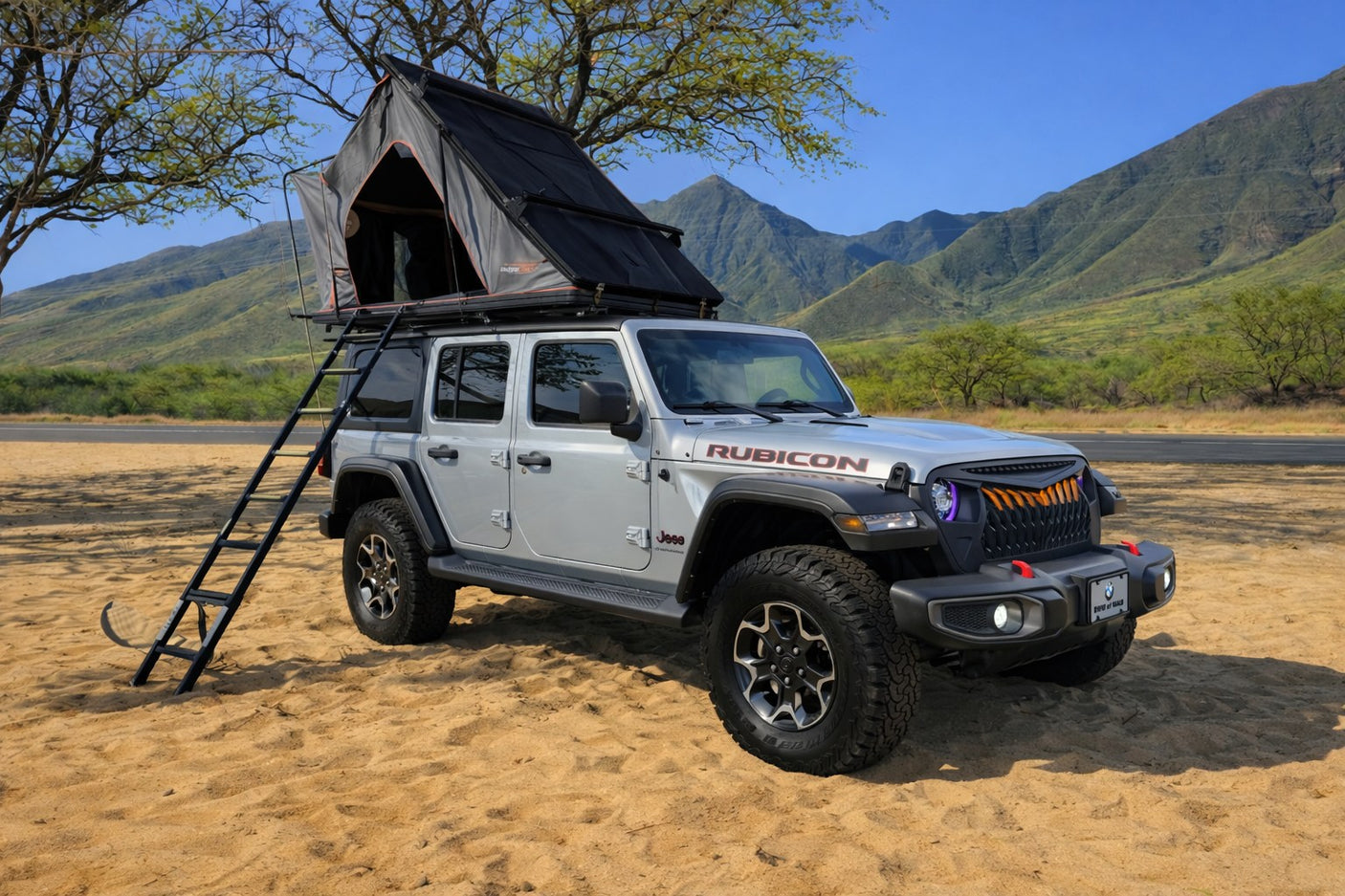 Silver Jeep Wrangler with rooftop tent & ladder on sand, mountains behind. Great rv rental for overlanding camping adventures!