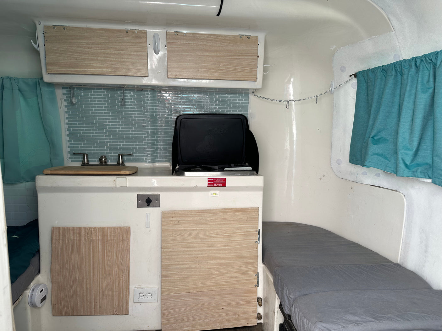 Compact RV rental interior with kitchen, light wood cabinets, tiled backsplash, and a grey cushioned bed area.