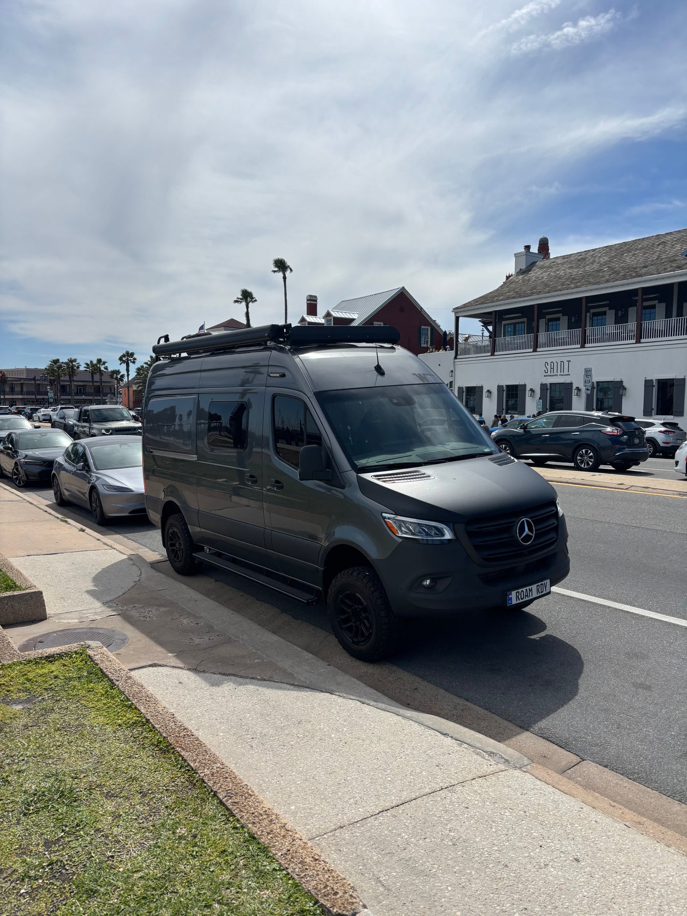 Dark gray Mercedes Sprinter RV rental, customized for travel, parked on a sunny street with buildings and palm trees.