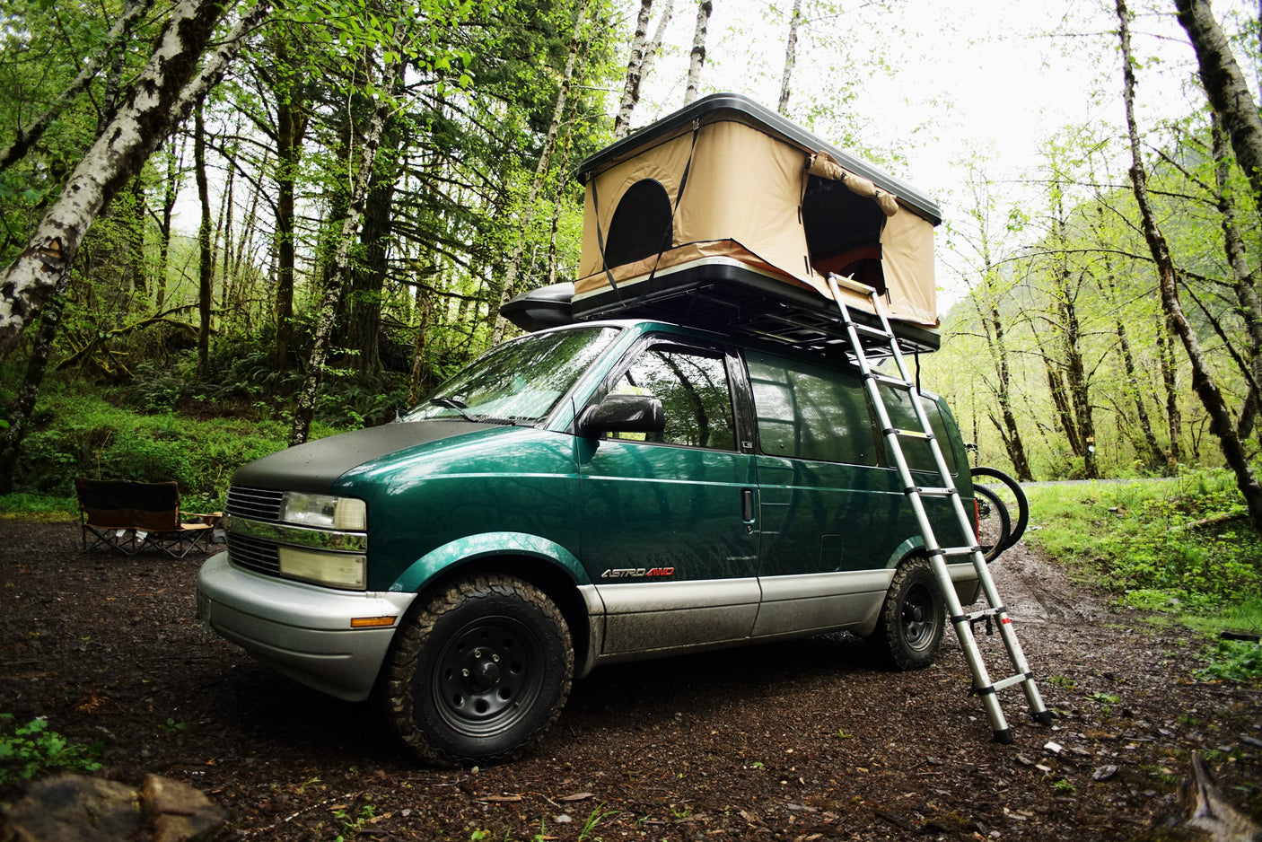 A green overlanding van with a rooftop tent and ladder parked on a dirt path in a lush forest. Perfect RV rental for immersive nature camping.