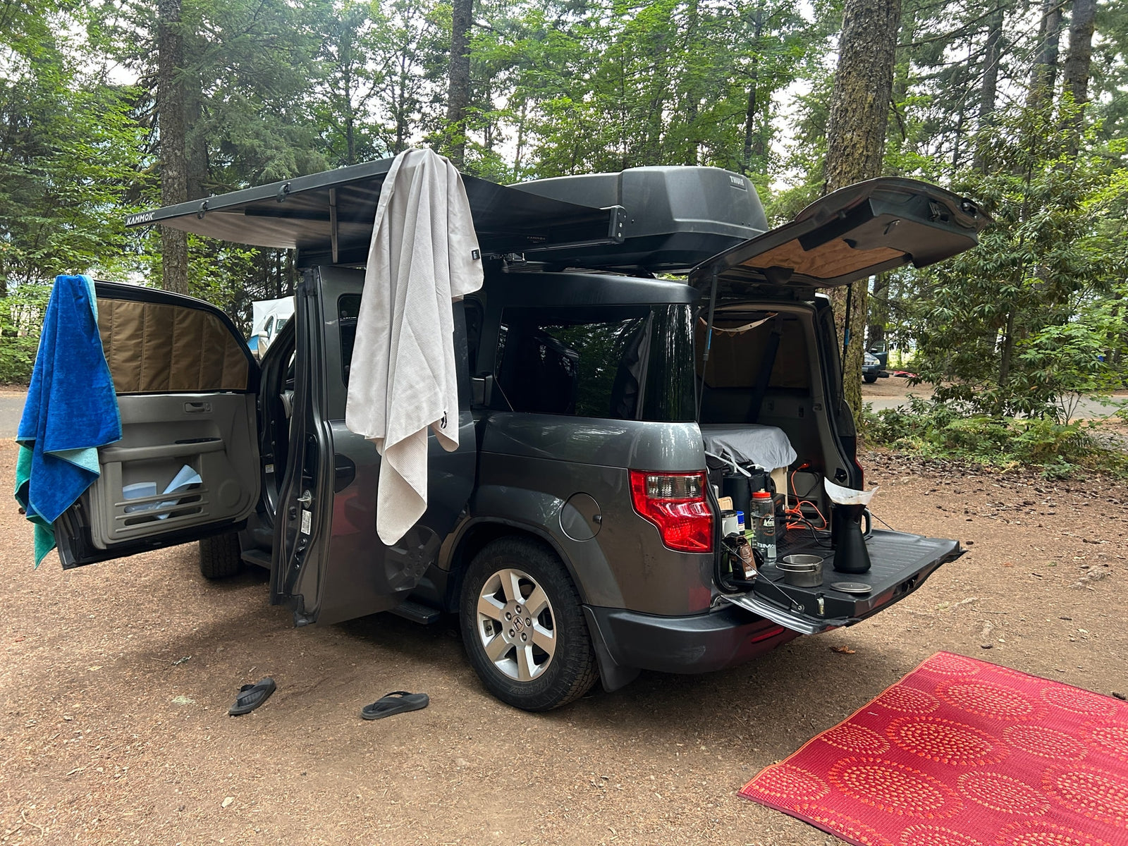 A grey Honda Element, set up like an RV rental for car camping, features an awning, cargo box, and a rear kitchen station.