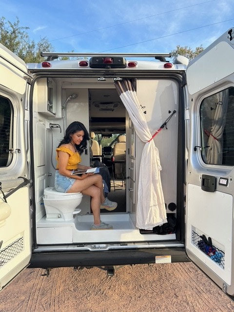 Enjoying a book in the compact bathroom of an RV rental, with open doors offering fresh air. Perfect for on-the-go comfort!
