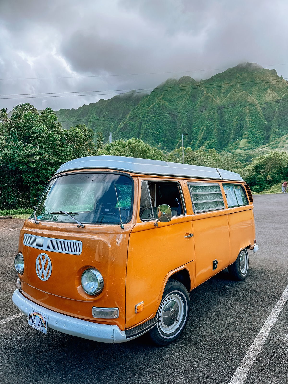 Orange vintage VW camper van (RV rental) parked in Hawaii, with lush green, cloud-covered mountains. — photo 4
