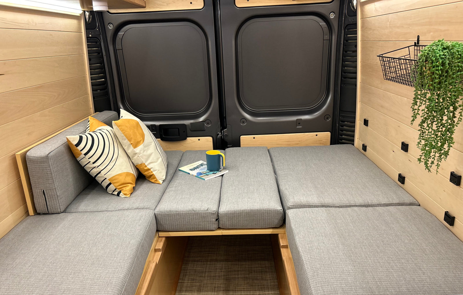 RV rental camper van interior featuring modular gray cushions that convert to a bed, light wood walls, and rear doors.