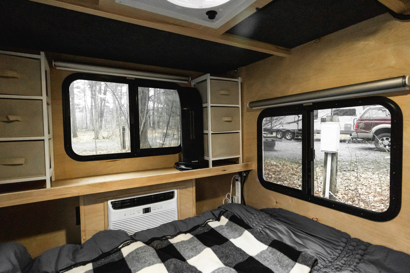 This RV rental interior features a cozy bed, AC unit, shelving, and two windows offering views of a wooded campsite.
