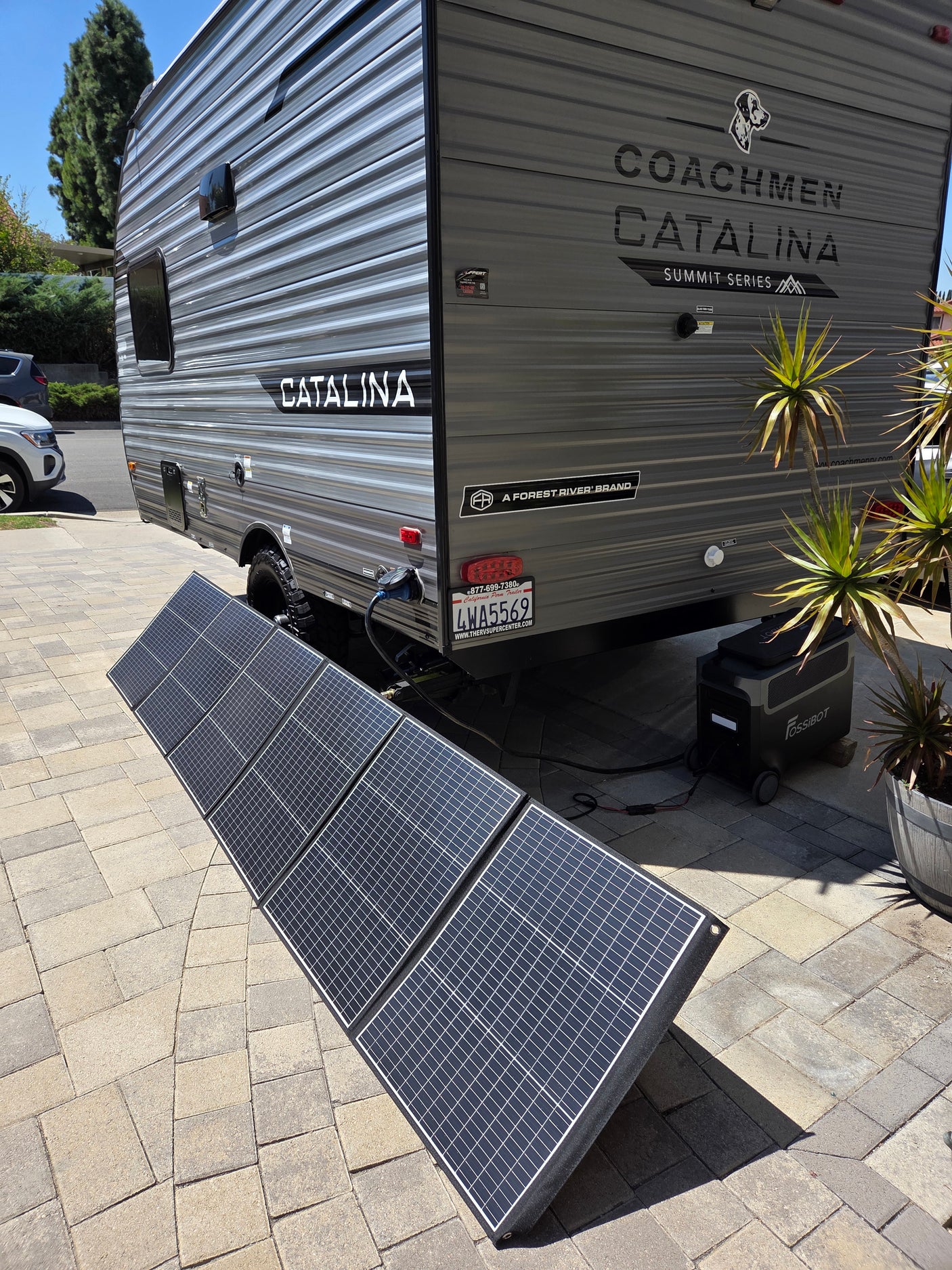 A gray Coachmen Catalina RV rental uses portable solar panels to charge a FOSSIBOT power station on a paved driveway.