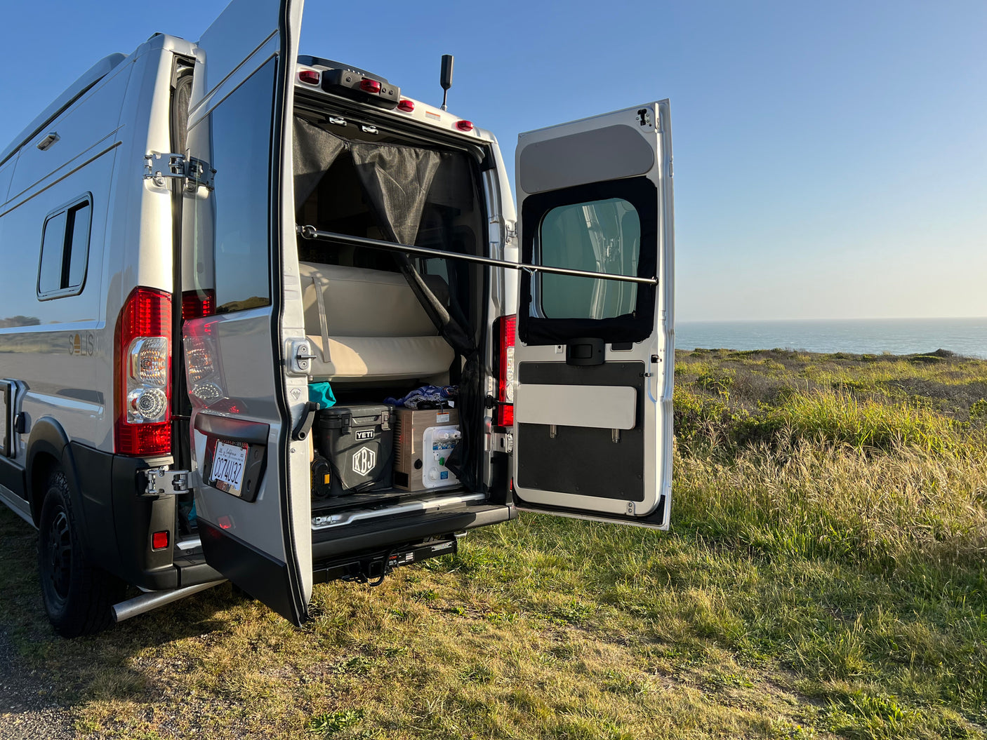 Silver RV rental camper van parked on a grassy cliff, back doors open revealing interior and ocean view. Enjoy scenic travels!