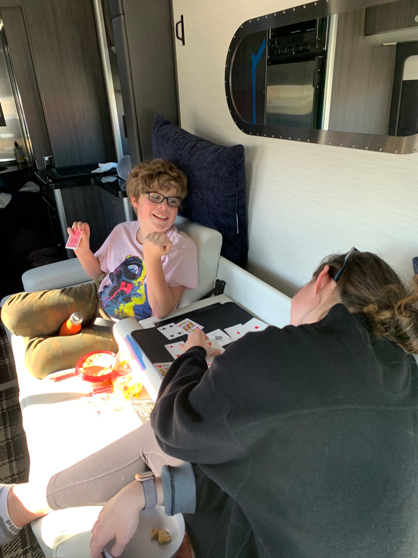 Two people enjoy a card game in a cozy RV rental. Snacks & a smiling boy with glasses are visible, perfect for travel.
