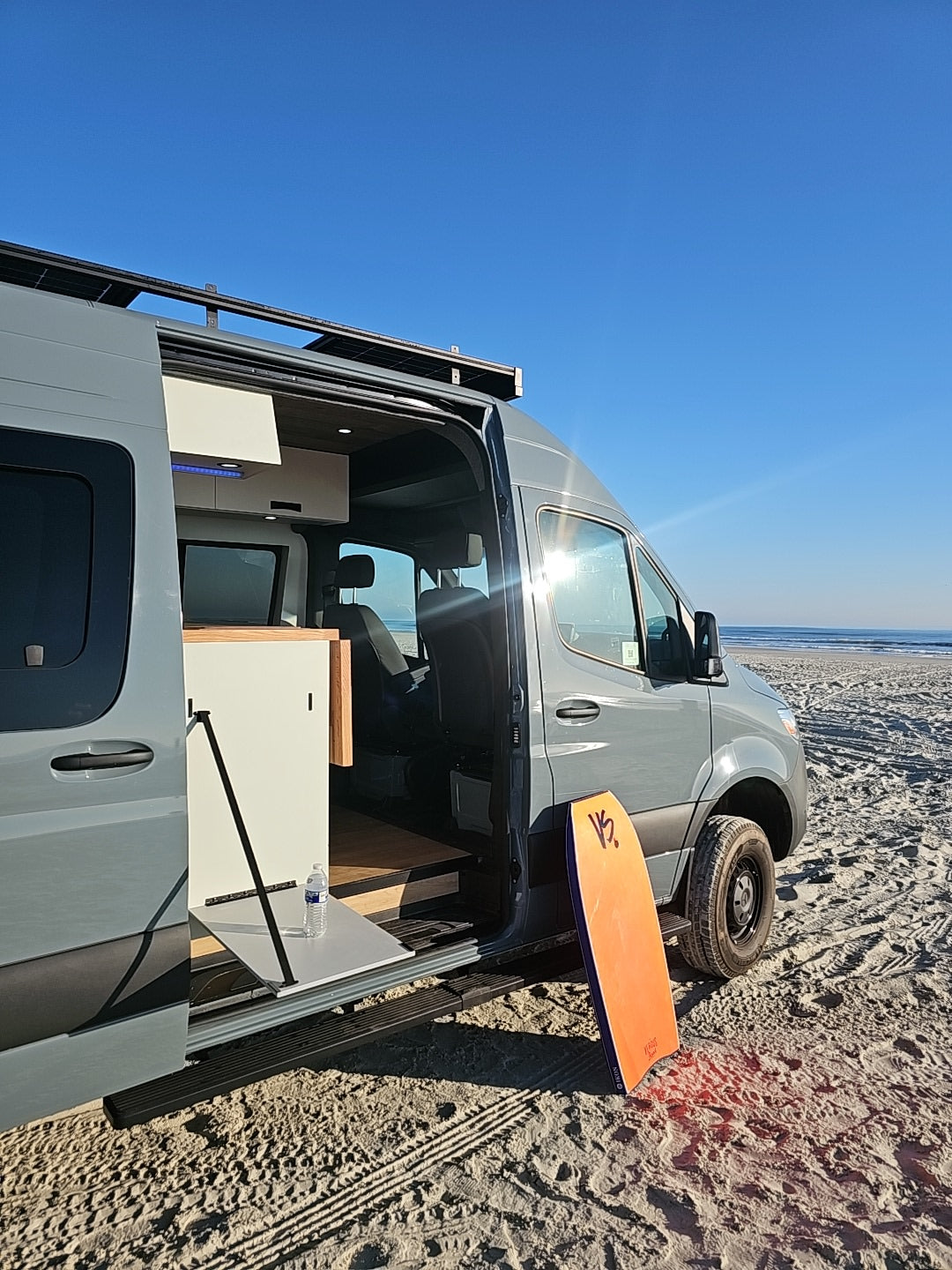 This RV rental van, ready for van life, is parked on a sunny beach. Door open, boogie board waiting for ocean fun.