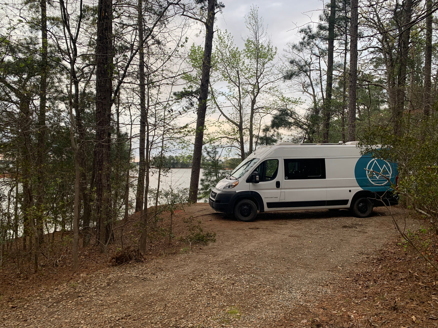 White camper van (RV rental) parked by a lake, surrounded by trees, suggesting a serene outdoor escape.