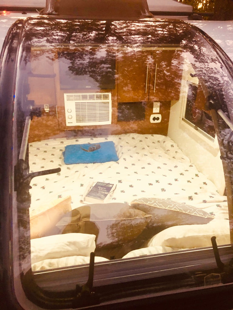 Cozy rv rental interior: bed, AC unit, and cabinets seen through its window. Reflections of trees are visible on the glass.
