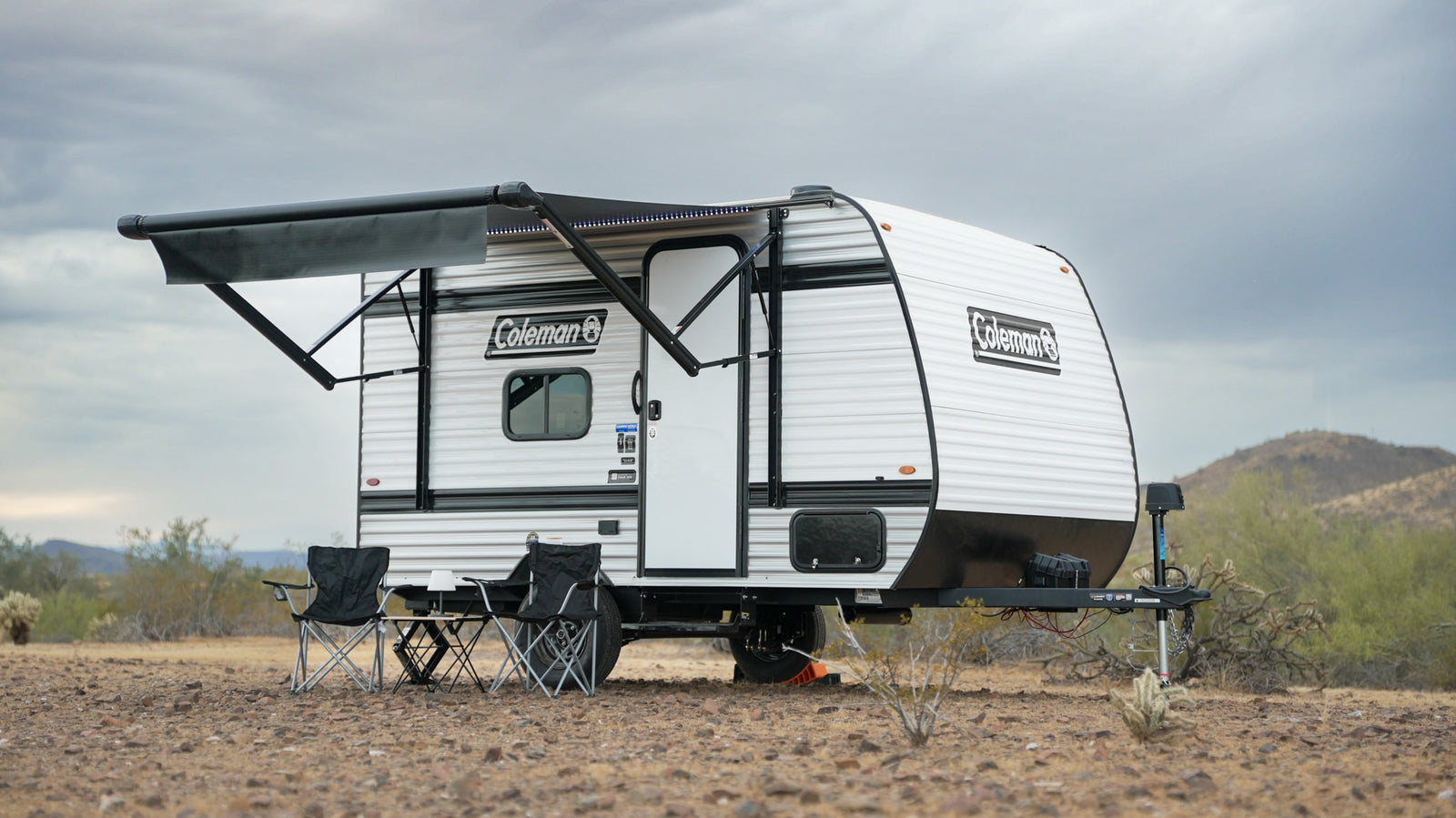 Coleman travel trailer rv rental with an extended awning and chairs set up in a sparse desert landscape under a cloudy sky.