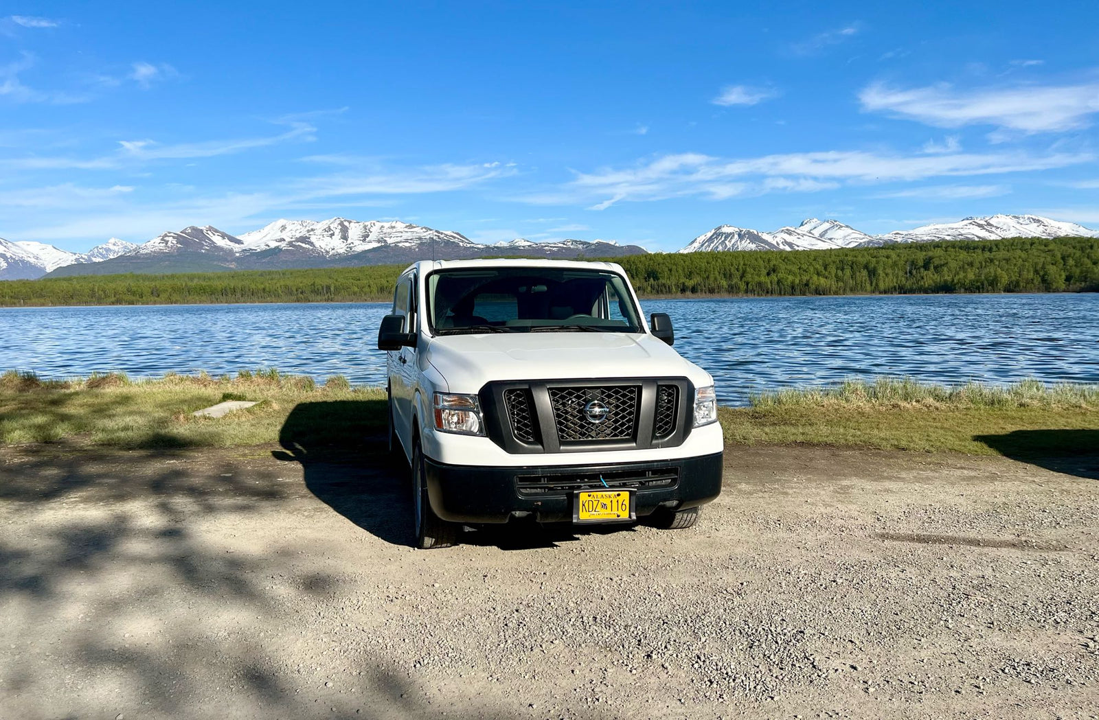 offering scenic views of snow-capped mountains under a blue sky.|White cargo van for an RV rental overlooking a scenic lake with mountains. A person is paddleboarding.|White rv rental van with Alaska plates facing a lake & snow-capped mountains under clear skies. Perfect for scenic travel.|Driver's view from an RV rental: spacious vehicle interior