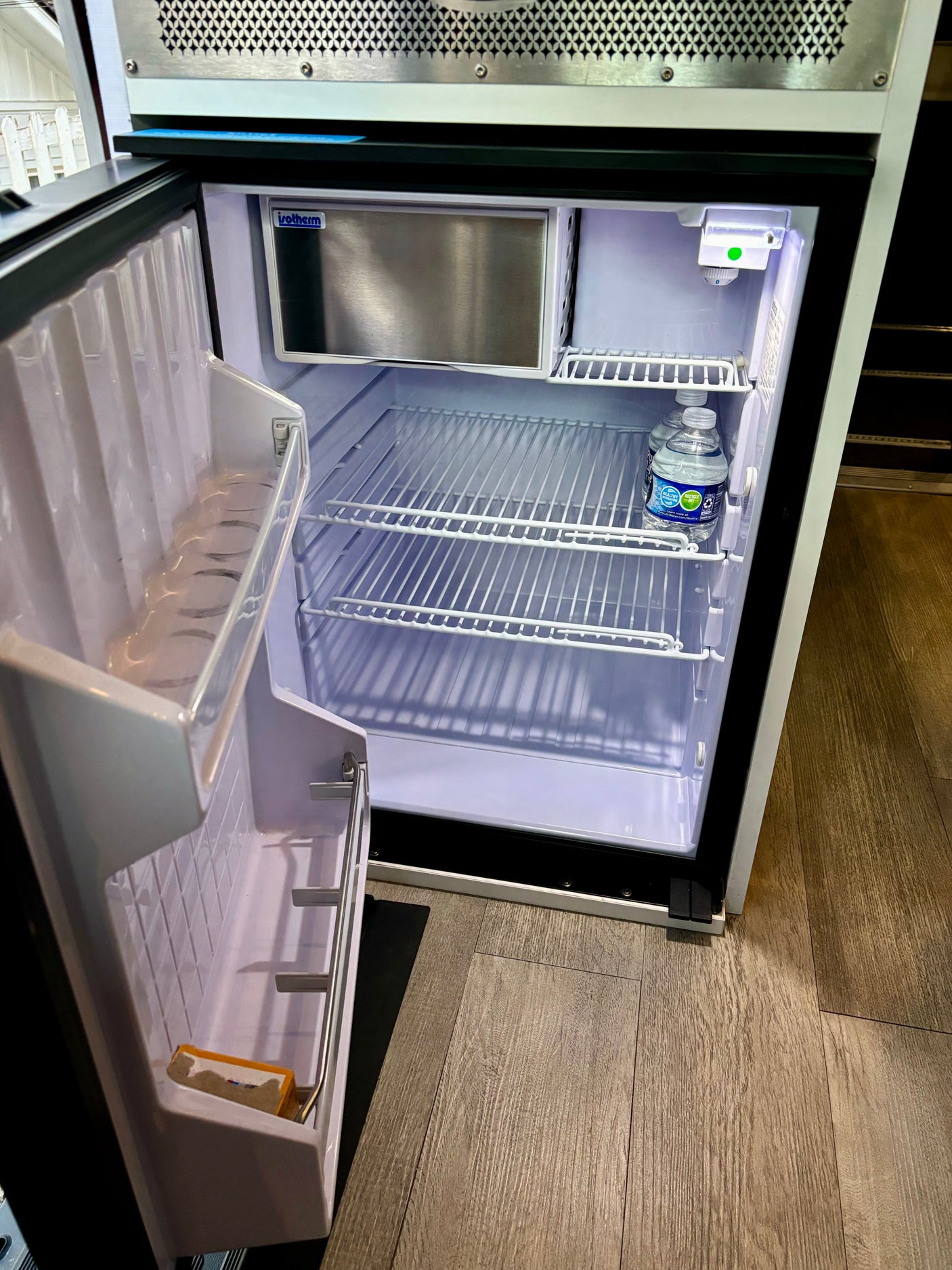 An open mini-refrigerator/freezer in an RV rental, showing wire shelves and two water bottles inside.