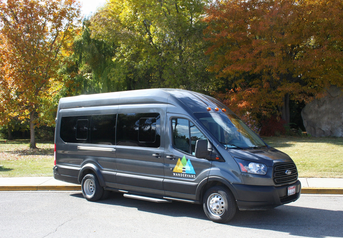 Grey WanderVans rv rental with tinted windows, parked on asphalt by a curb, surrounded by colorful fall trees.