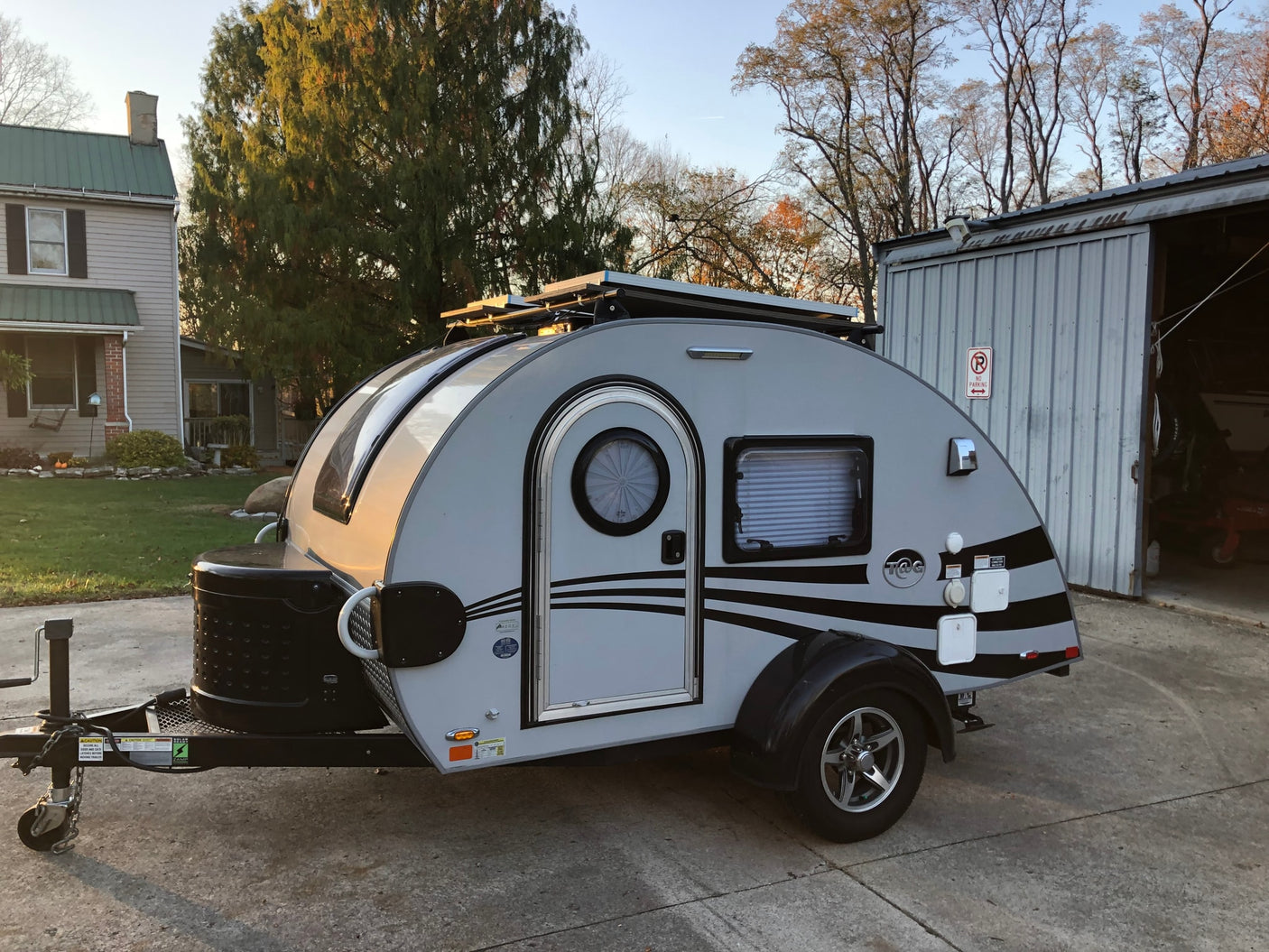 Compact grey teardrop RV rental with solar panels, parked on a driveway by a house and shed, ready for adventure.