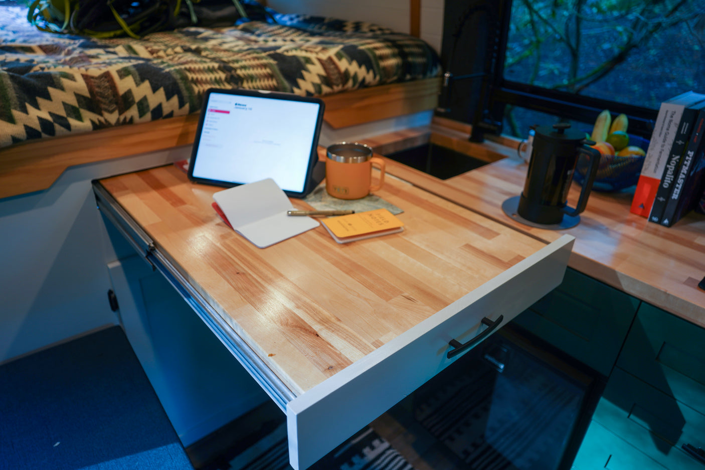 Inside an RV rental: a compact, modern interior with a pull-out wooden desk, tablet, bed, and kitchenette.