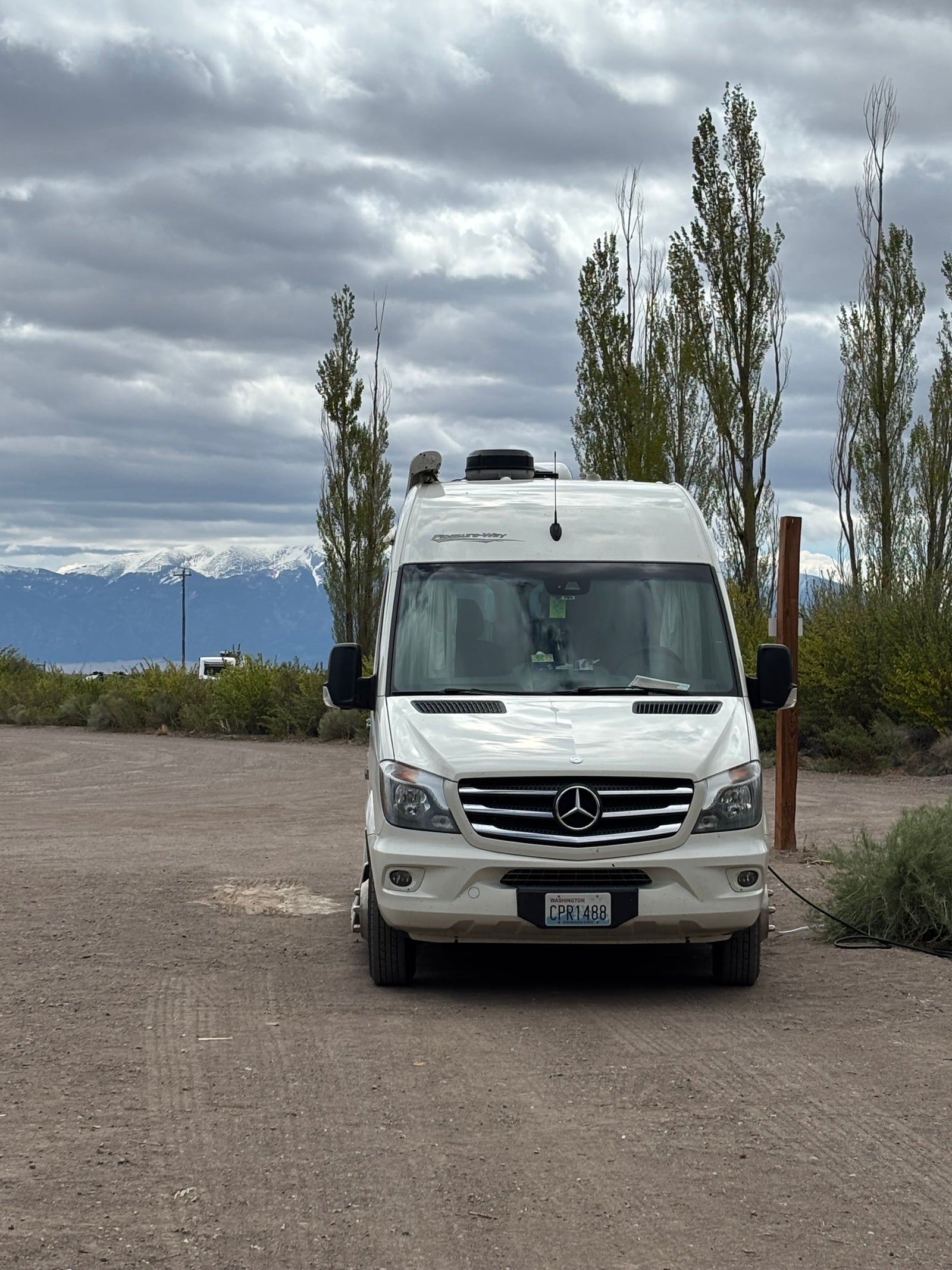 White Mercedes Sprinter RV rental parked on gravel, with snow-capped mountains and tall trees under cloudy skies.