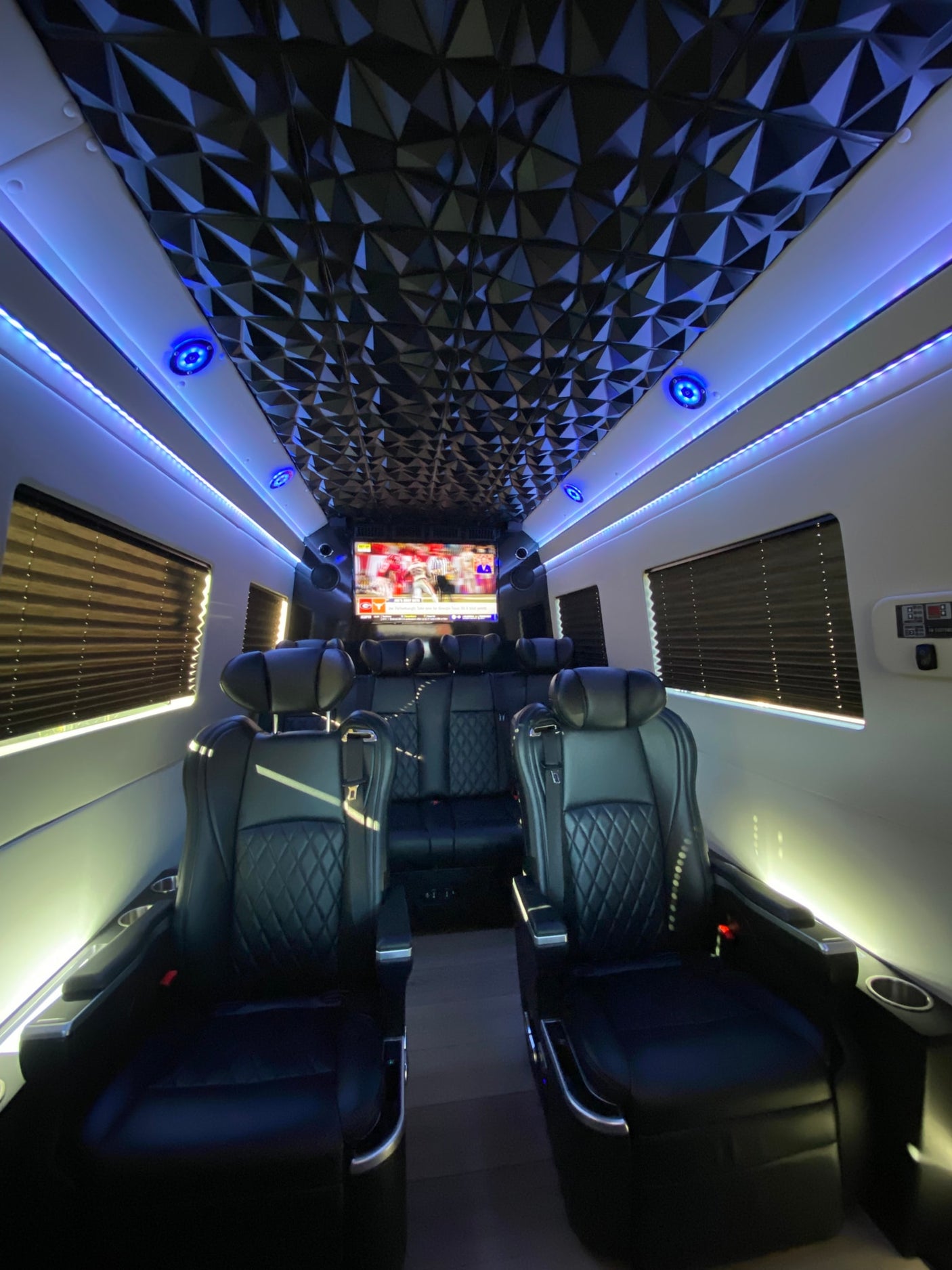 A plush RV rental interior features black quilted captain chairs, a large screen playing sports, geometric ceiling, and blue accent lighting.