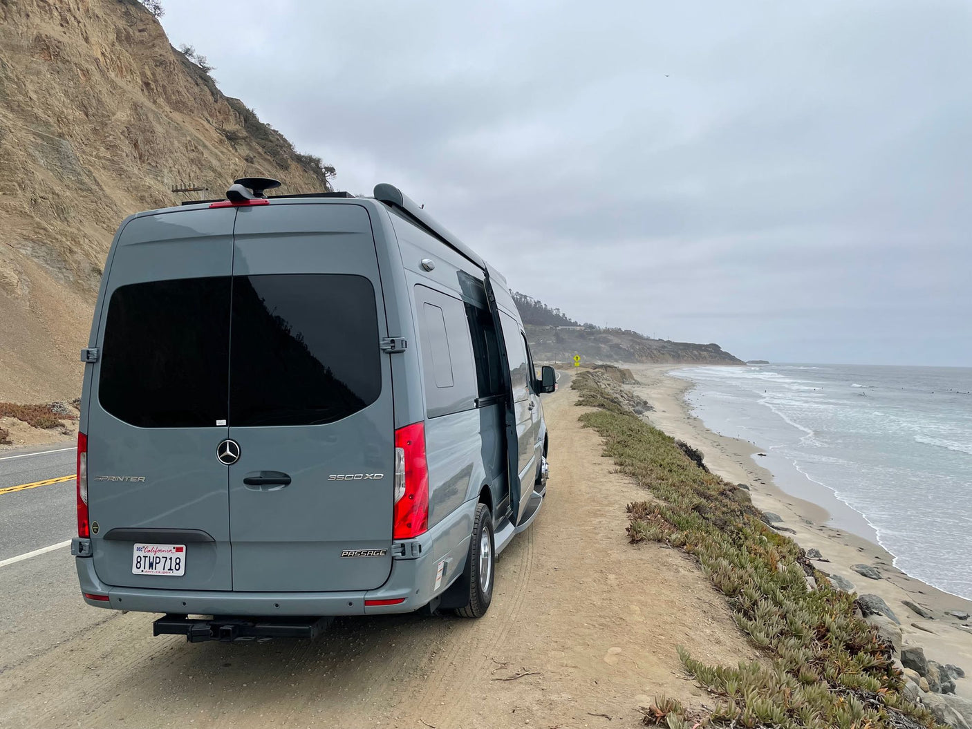Gray Mercedes Sprinter **rv rental** parked on a California coastal road by the beach and cliffs under an overcast sky. Perfect adventure.