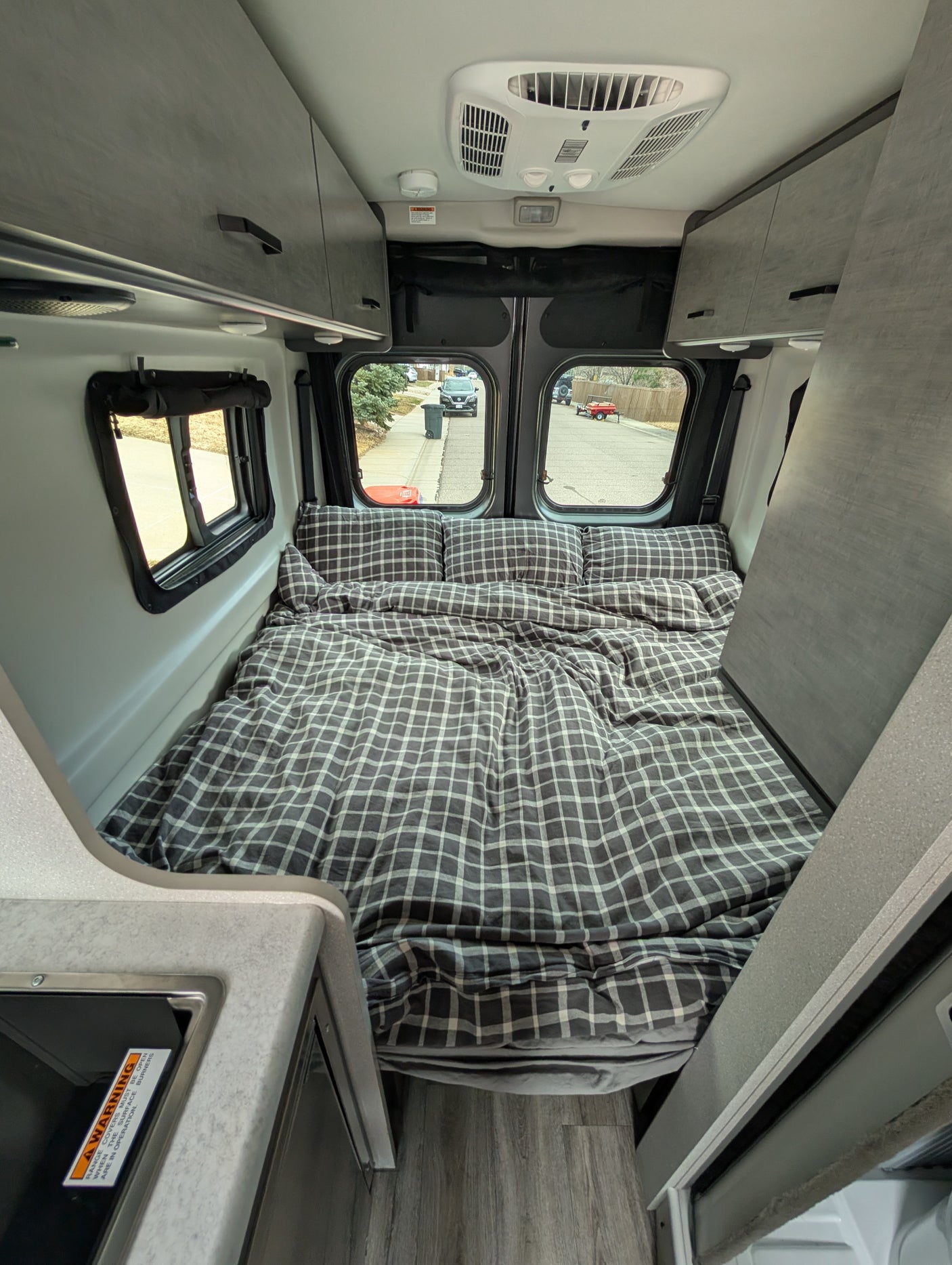 Inside a comfy RV rental: A grey plaid bed, overhead cabinets, and compact kitchen area for mobile travel.