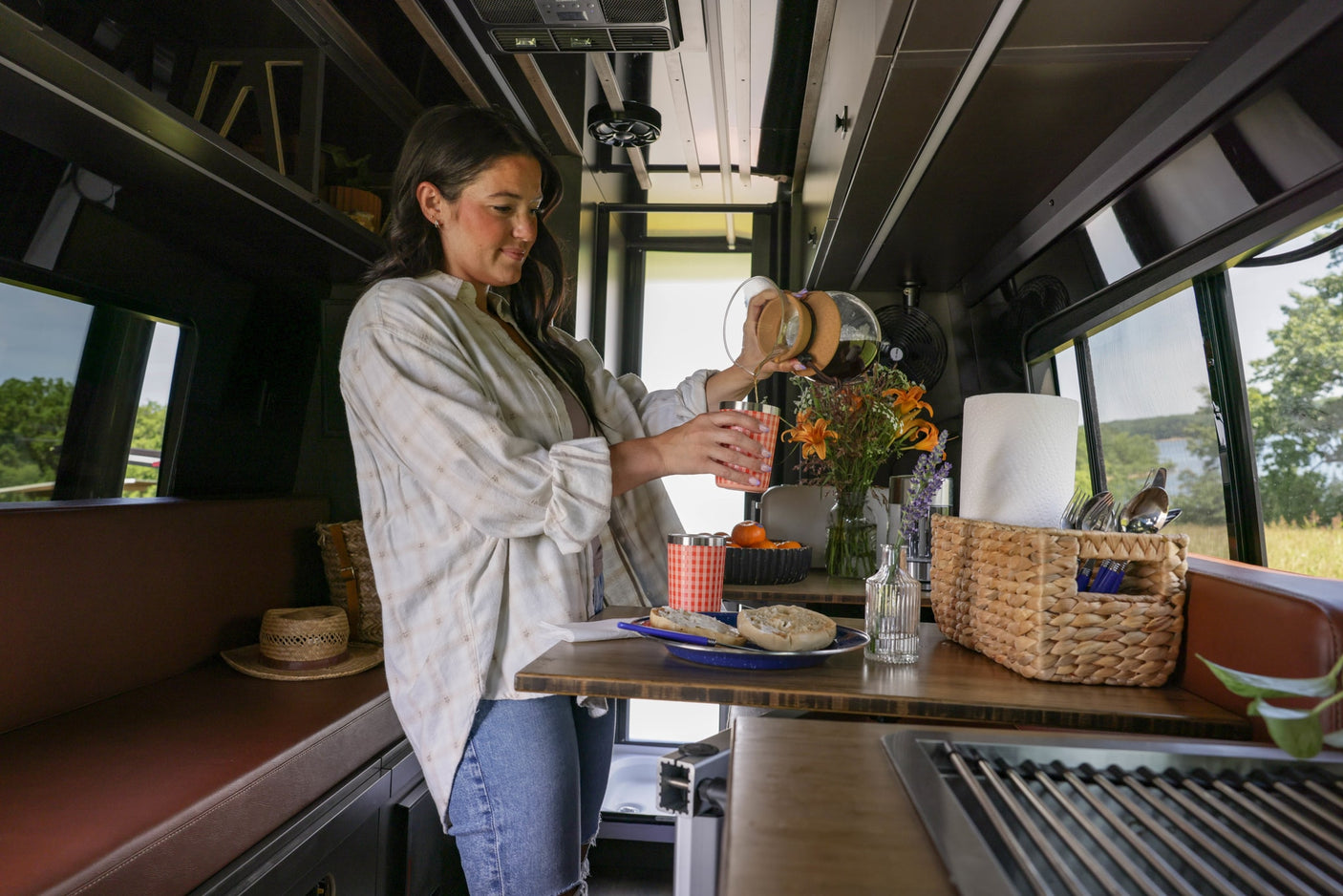 Woman pours coffee in her RV rental's mobile kitchen. Enjoying breakfast with bagels & a scenic nature view through the window.