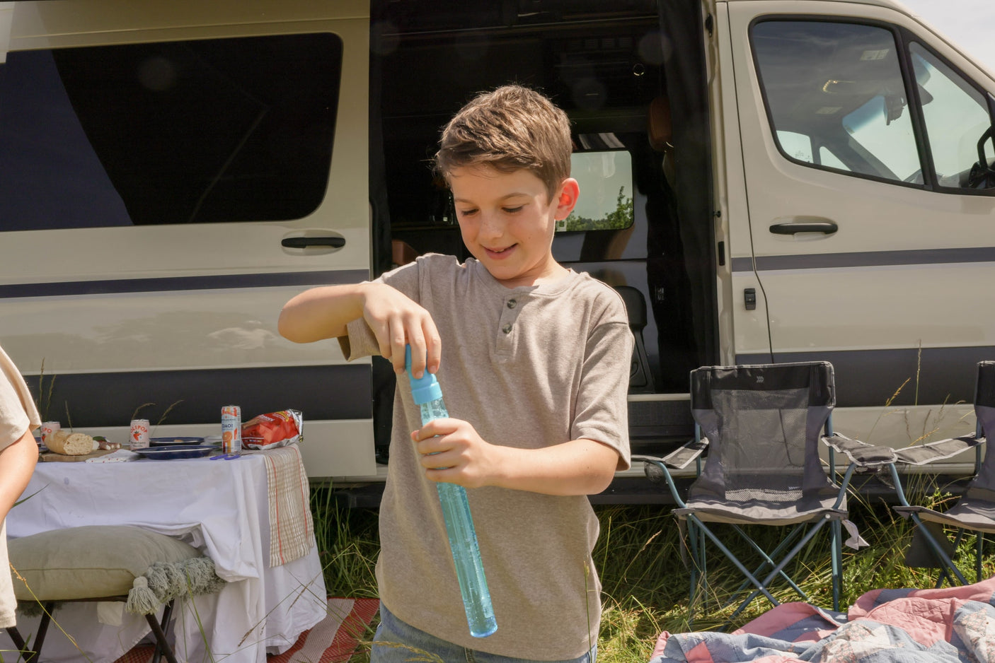 A smiling boy holds a blue bubble wand next to a light-colored RV rental with a picnic table and chairs.