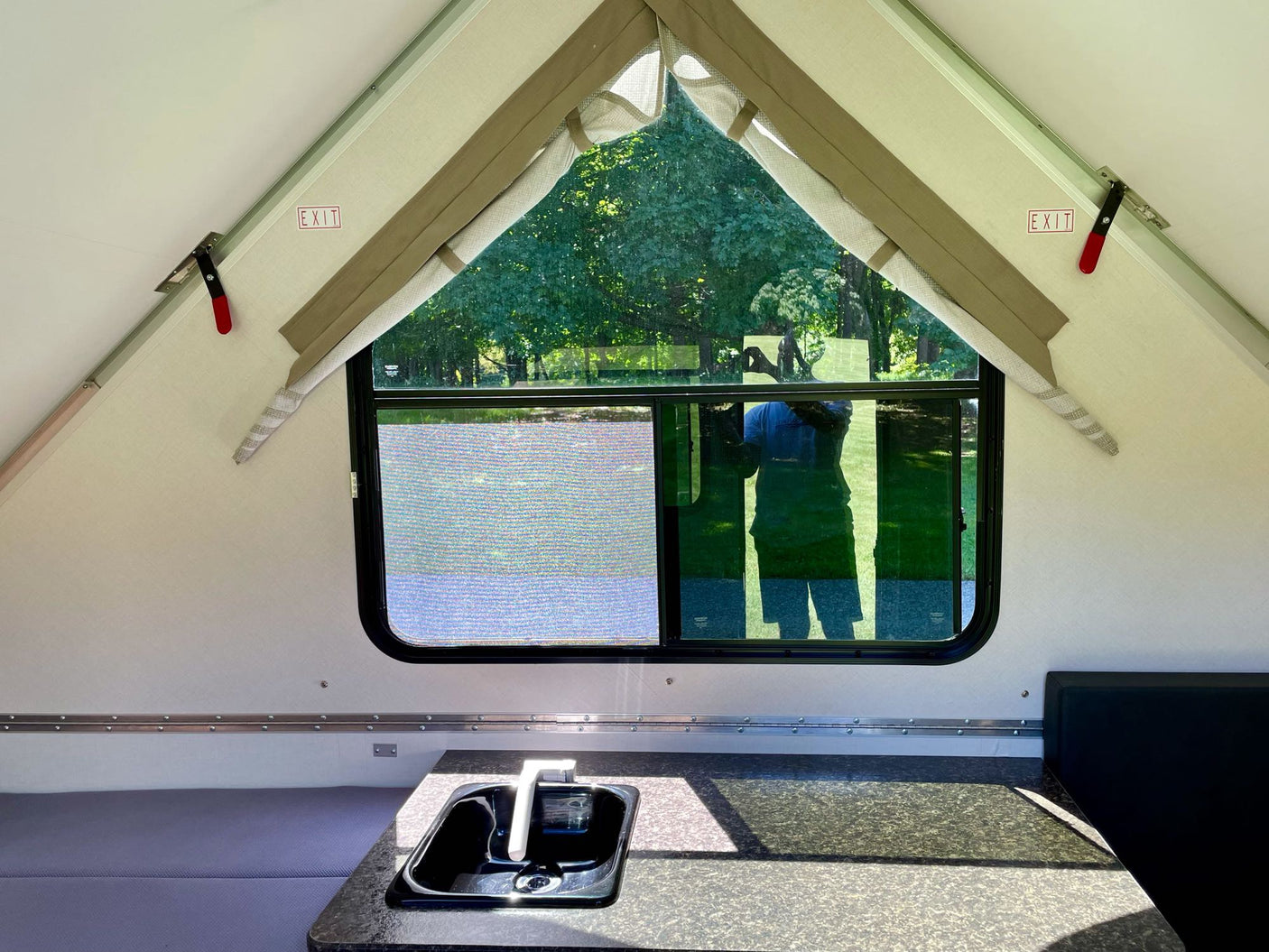 An RV rental A-frame camper interior featuring a sink, bed, and a large window overlooking a forest.