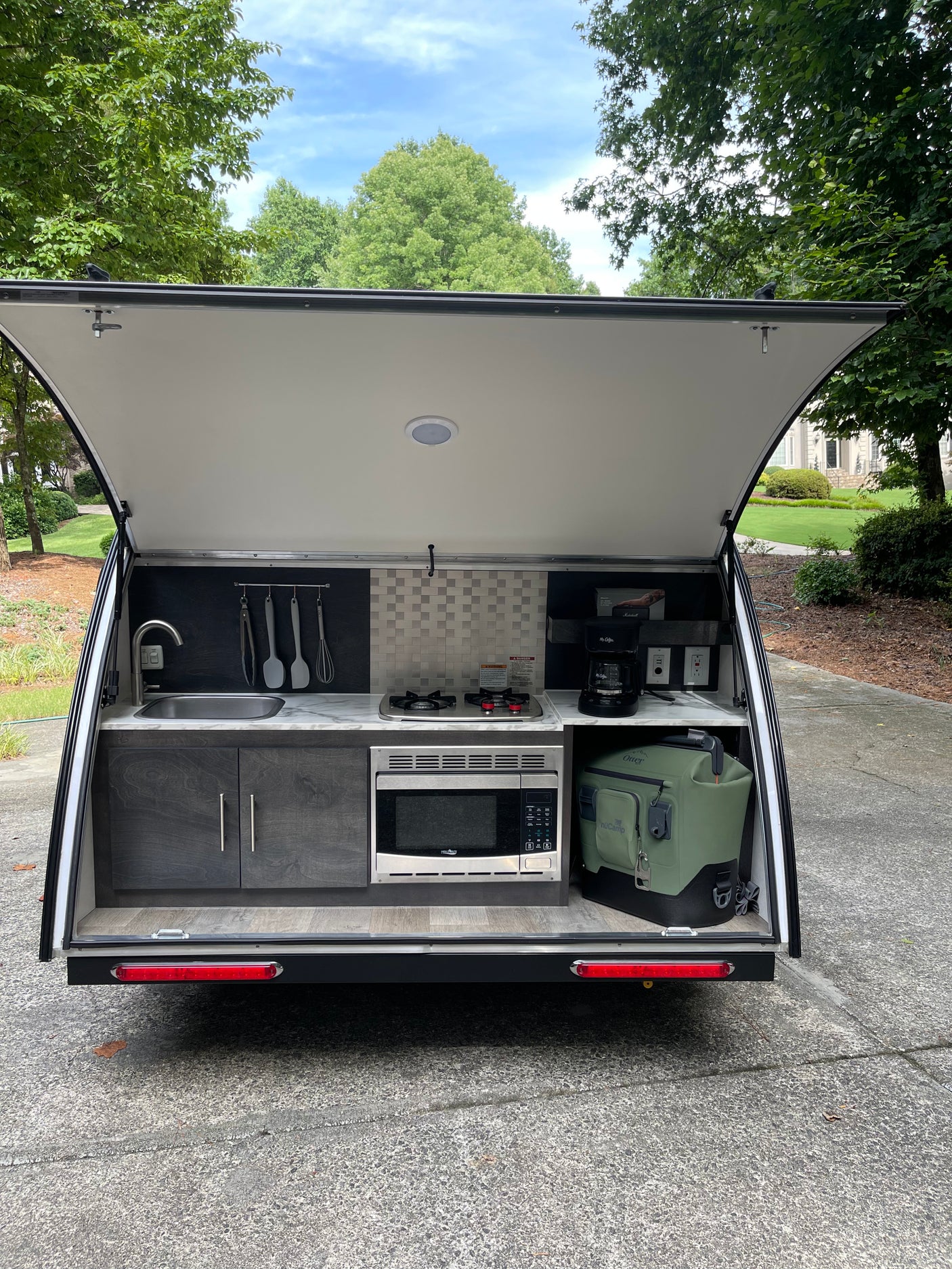 This rv rental features an open-hatch outdoor kitchen with a sink, stovetop, microwave, coffee maker, and cooler setup.