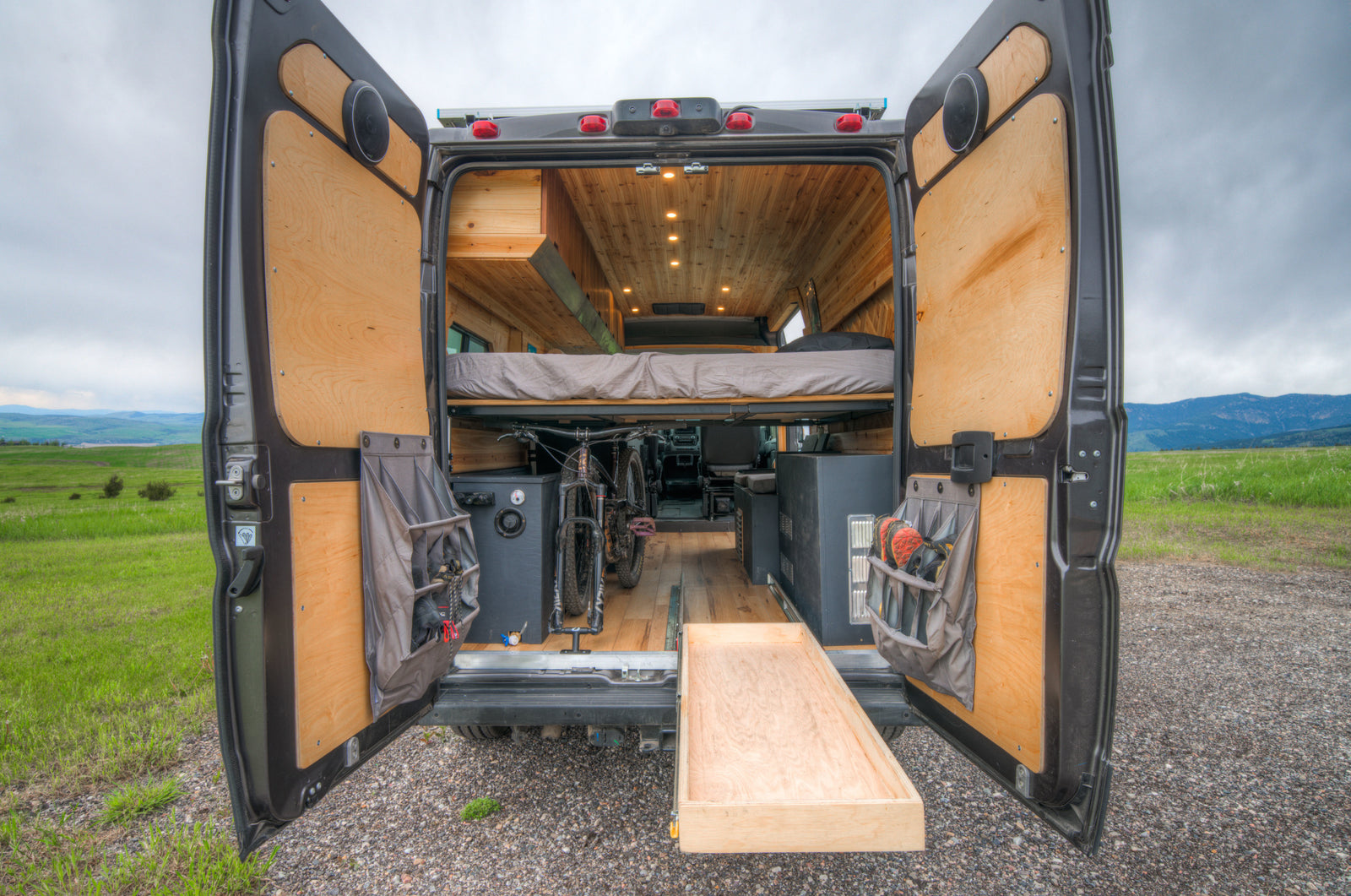 RV rental: Rear view of a custom camper van, doors open, revealing a wood interior, bed, bike storage & pull-out tray in nature.
