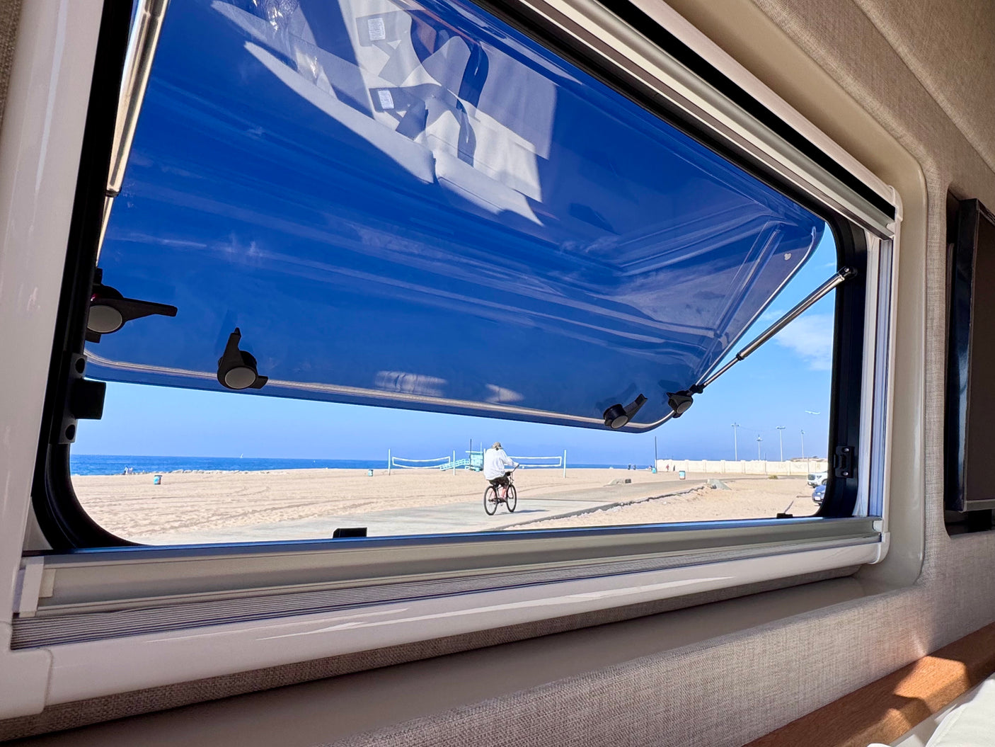 Perfect RV rental view! An open RV window overlooks a sunny beach, with a cyclist on a paved path and the ocean beyond.