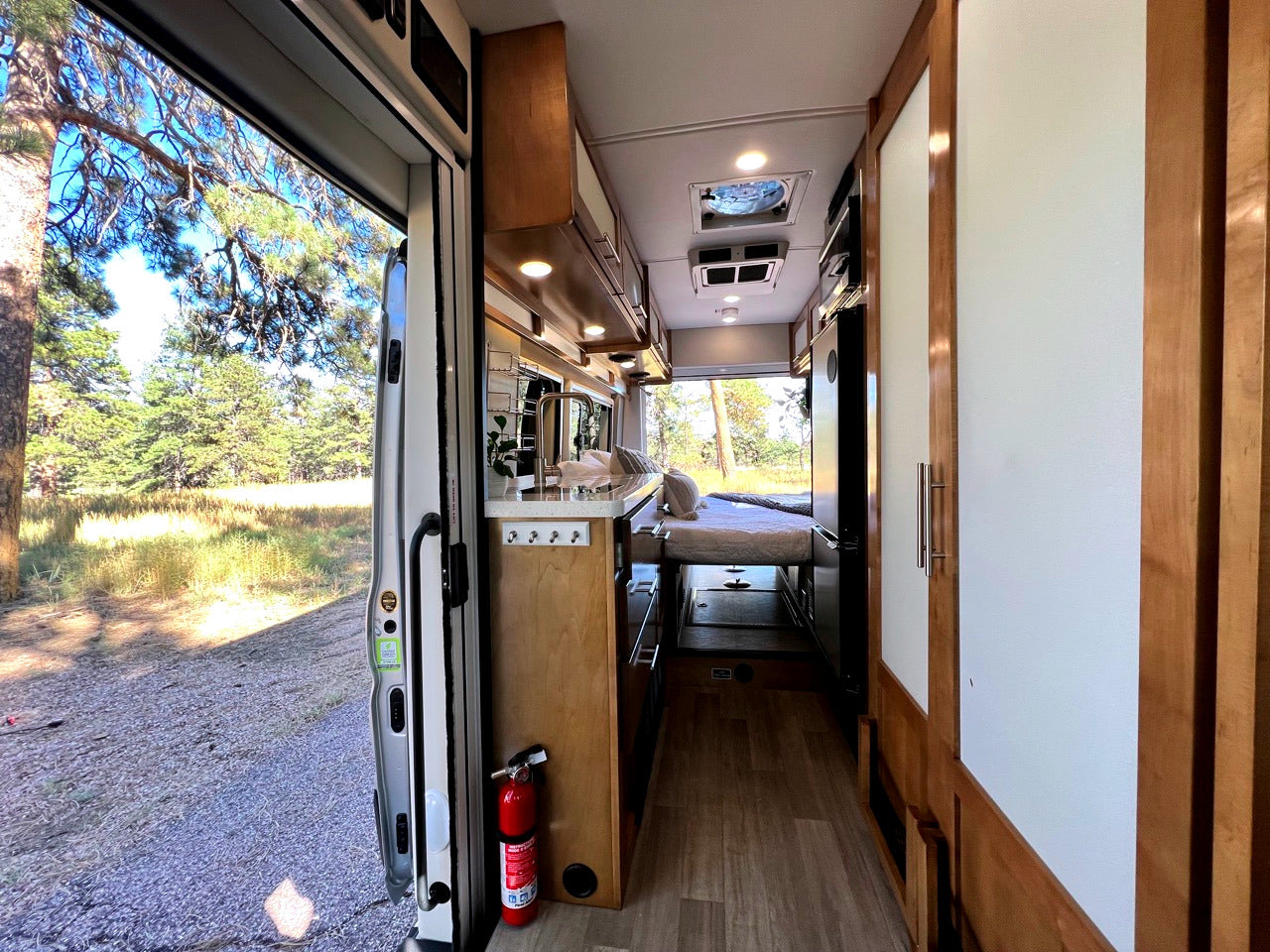 Spacious RV rental interior with kitchen, bed, and open door revealing a serene pine forest. Ready for adventure!