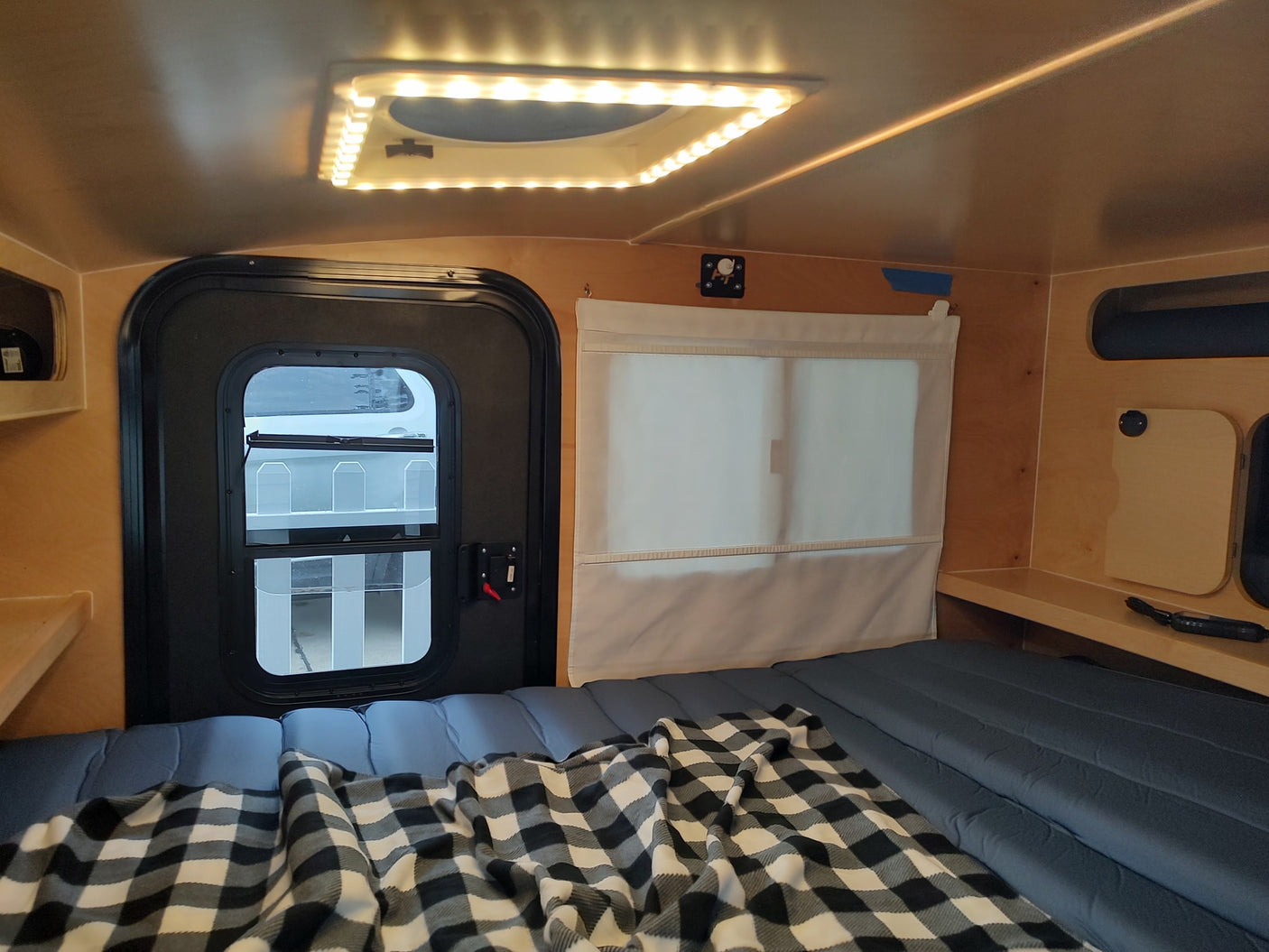 Compact RV rental interior featuring a comfortable bed with plaid blanket, a door, window with shade, and overhead LED lighting.